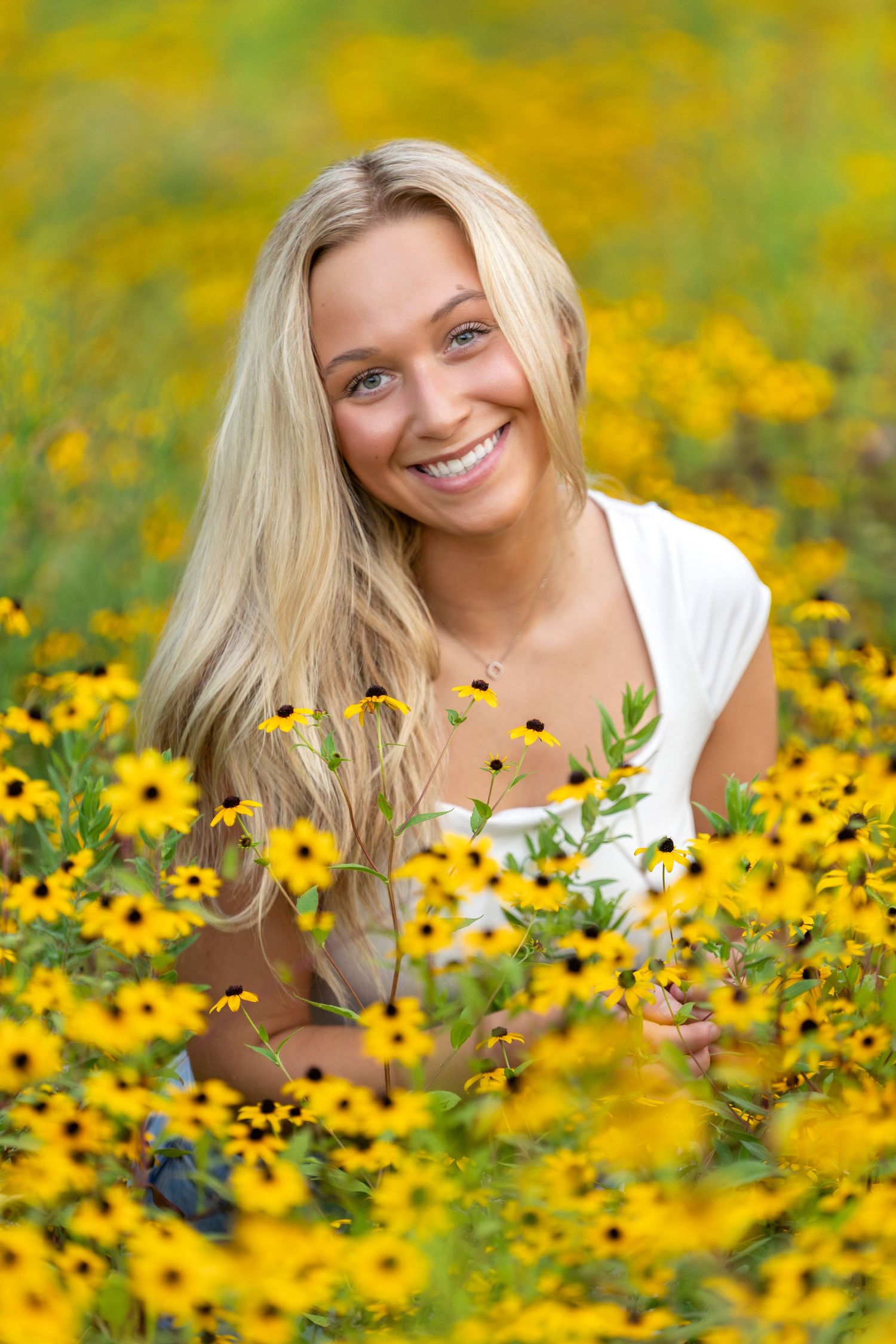 Smiling blonde woman sitting in a vibrant field of yellow black-eyed Susan flowers on a sunny day.