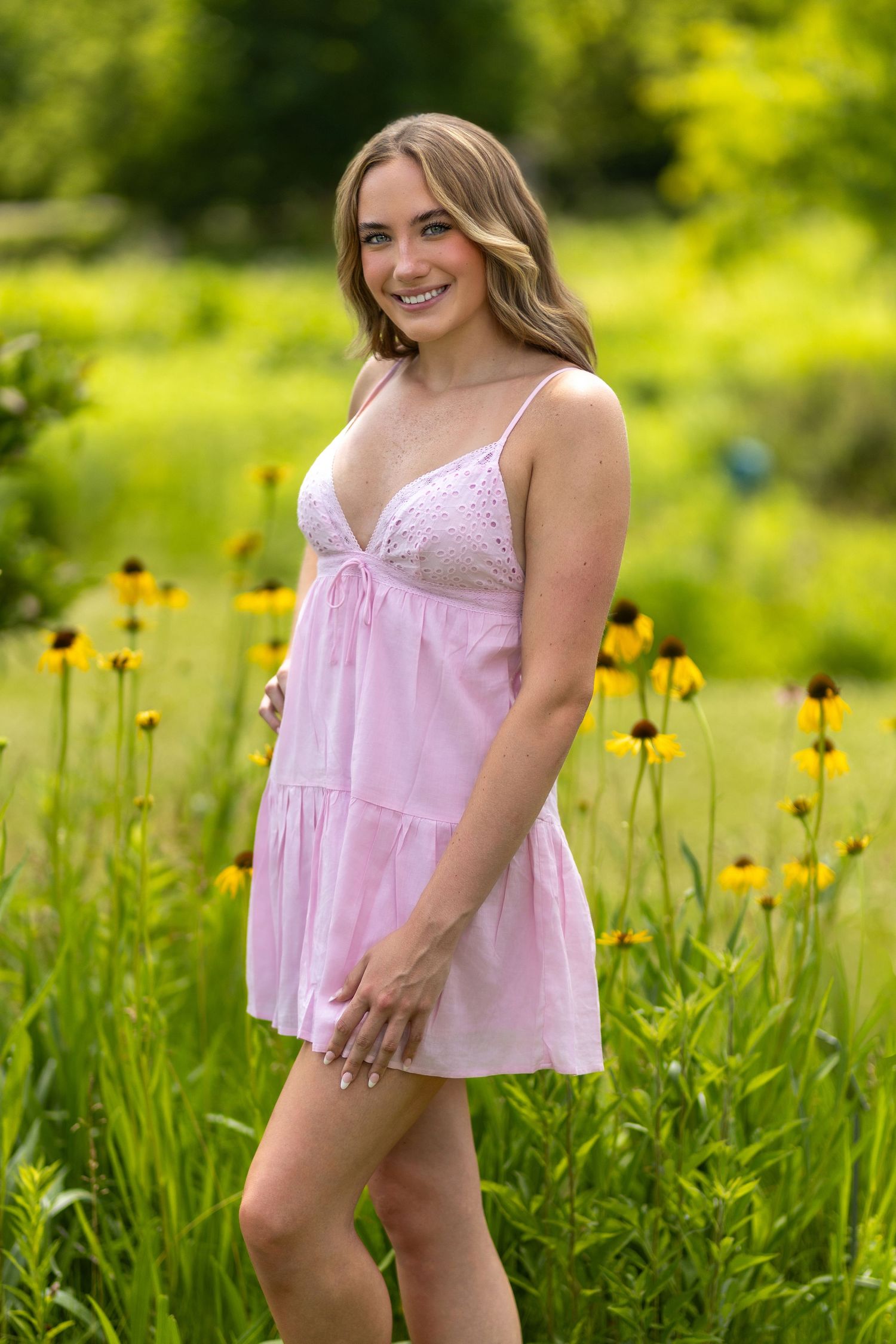Young woman in pink mini dress smiling in a field of yellow wildflowers on a sunny day.