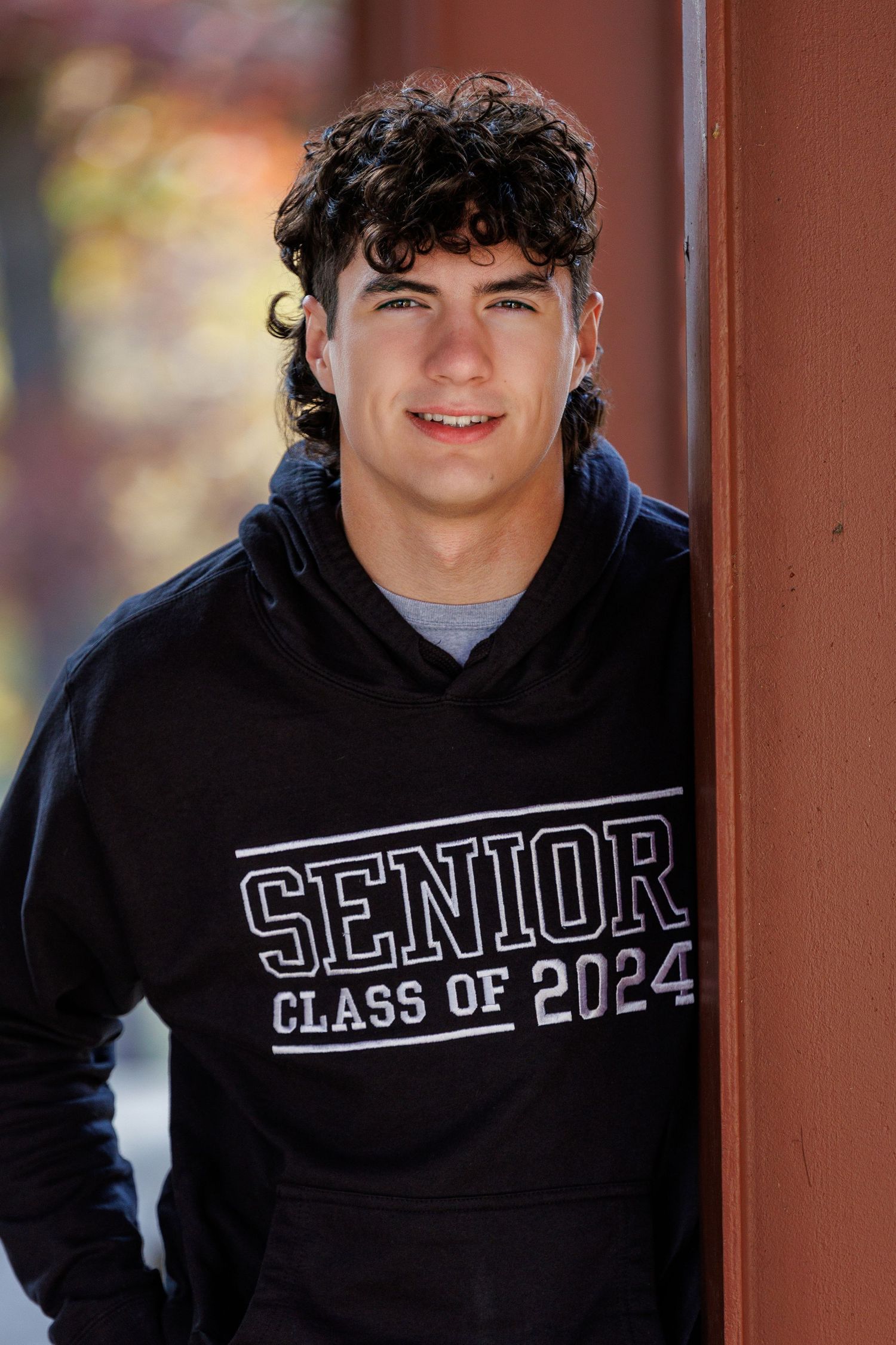 Smiling high school senior wearing a black Class of 2024 hoodie, leaning against a wooden post outdoors.