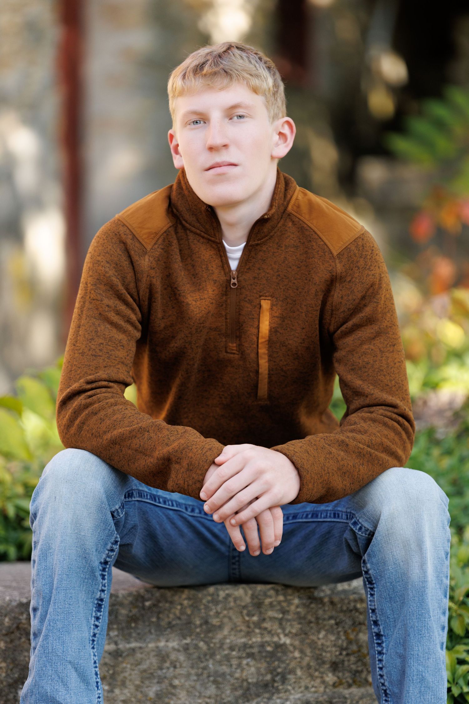 Young man with blonde hair sits outdoors in brown zip-up sweater and jeans, posing for a portrait photo.