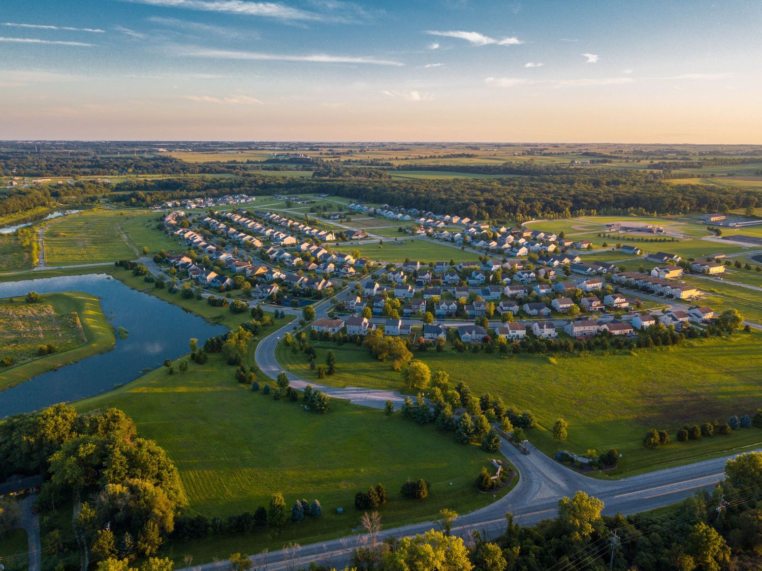 Aerial view of a suburban neighborhood with curved streets, a pond, and lush green landscapes at golden hour.