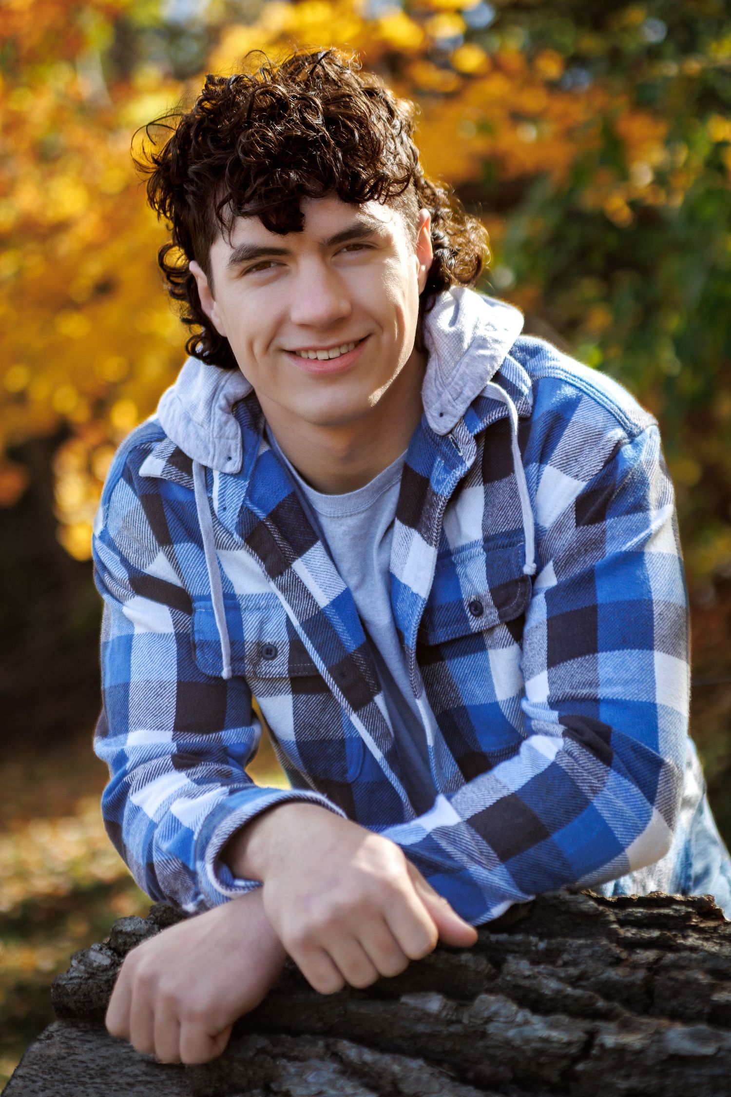 Young man with curly dark hair smiling outdoors, wearing a blue plaid hoodie shirt, leaning on a log with autumn foliage.