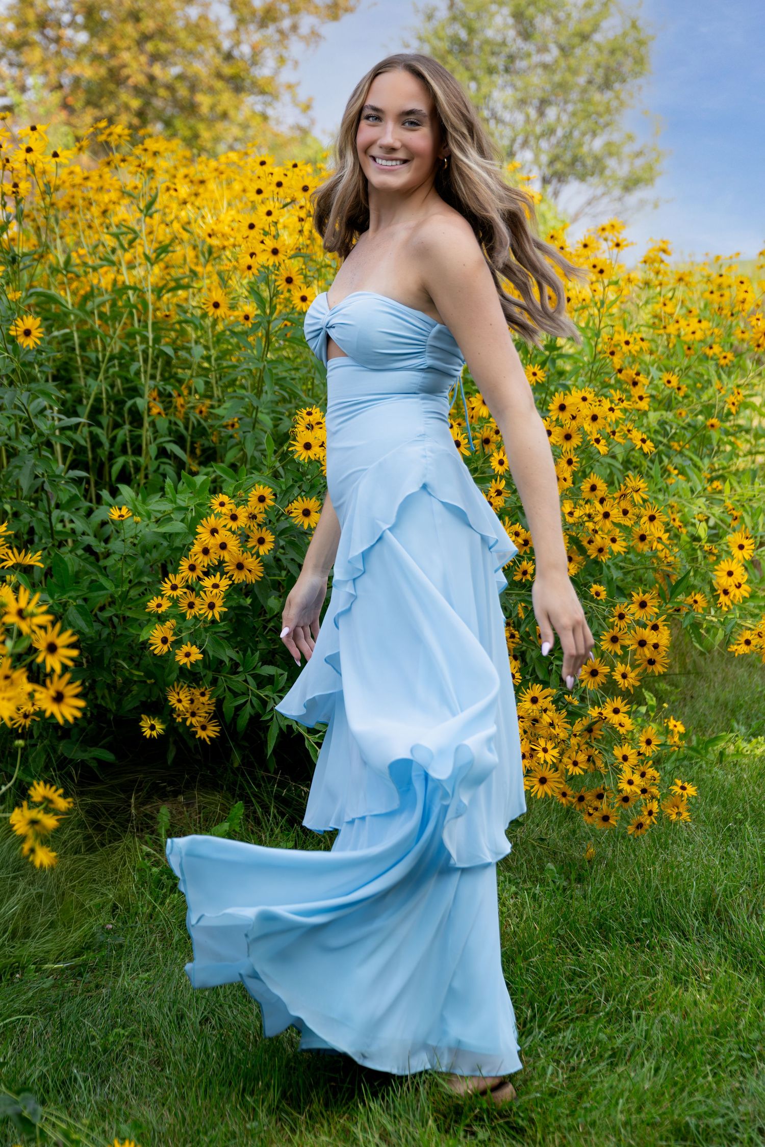 Young woman in flowing light blue strapless gown posing among vibrant yellow wildflowers outdoors.