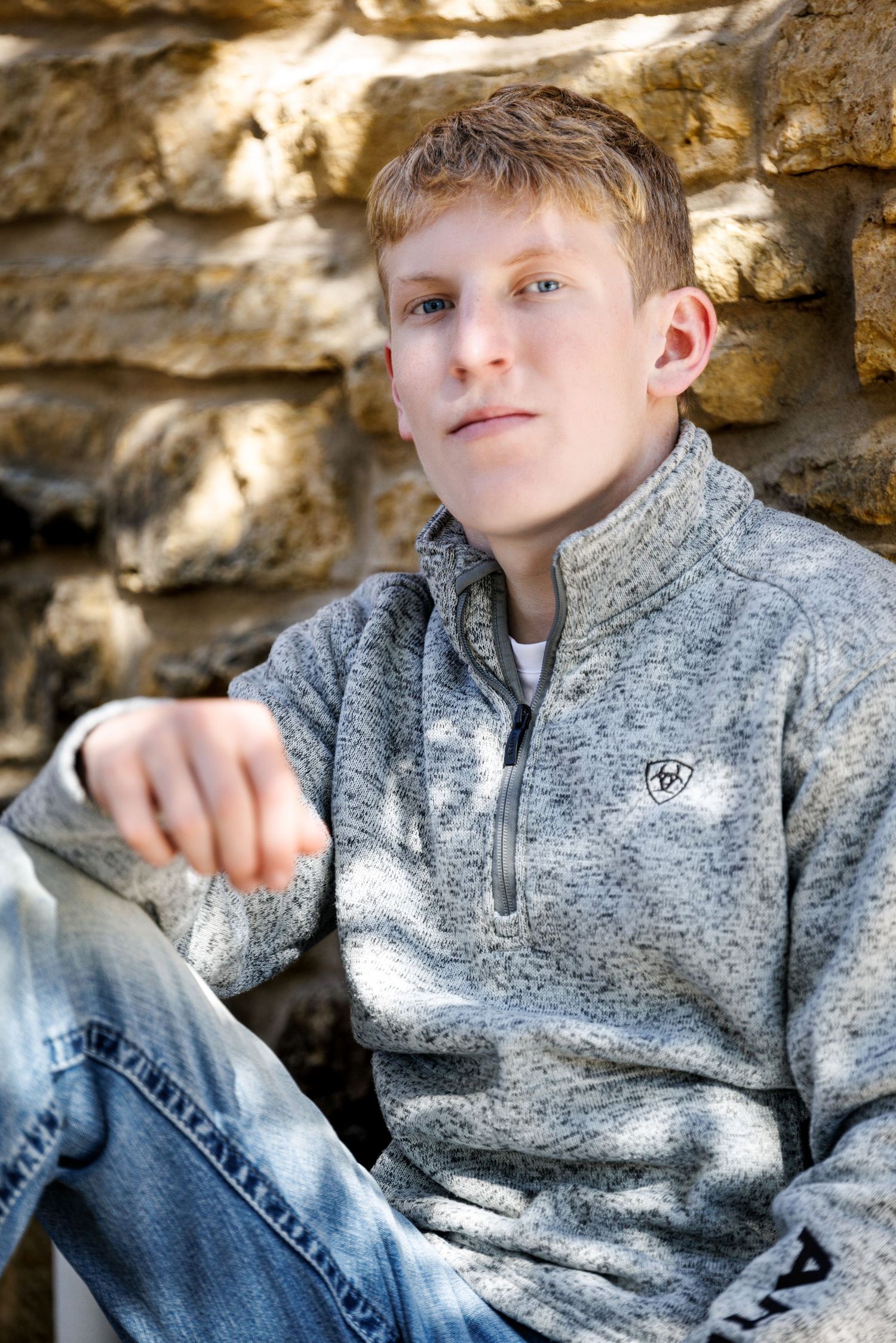 A teenage boy in a gray Ariat quarter-zip sits casually against a stone wall, wearing jeans, with a relaxed expression.