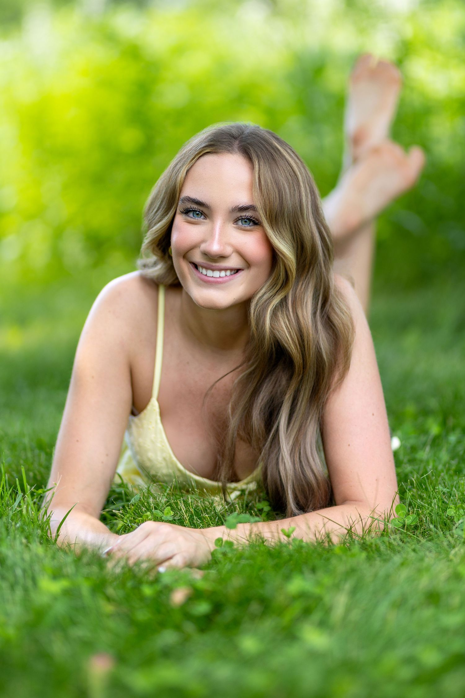 Young woman with long blonde hair smiling while lying on green grass outdoors in summer.