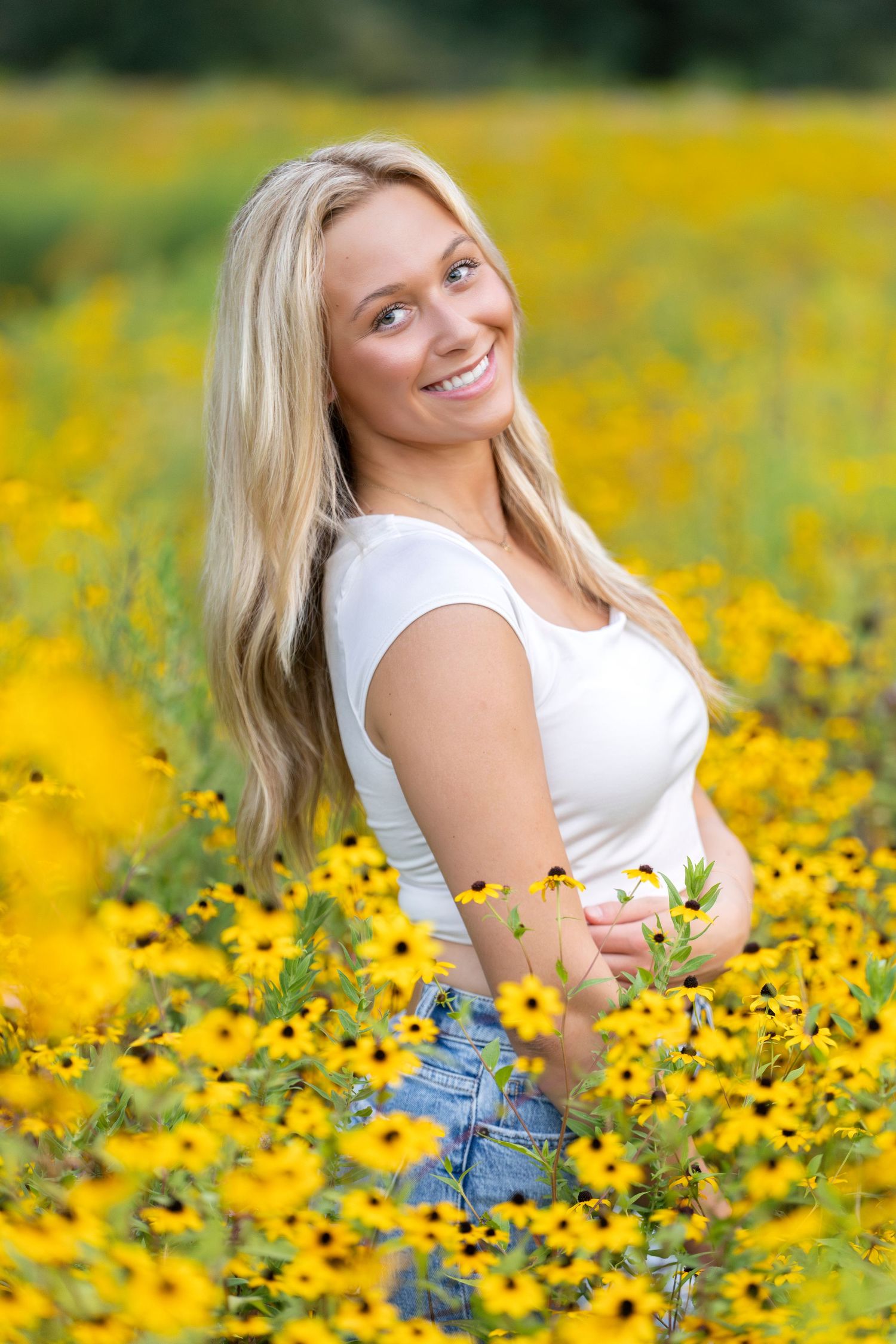 Smiling blonde woman in white top sitting among vibrant yellow black-eyed Susan flowers in a sunny field.