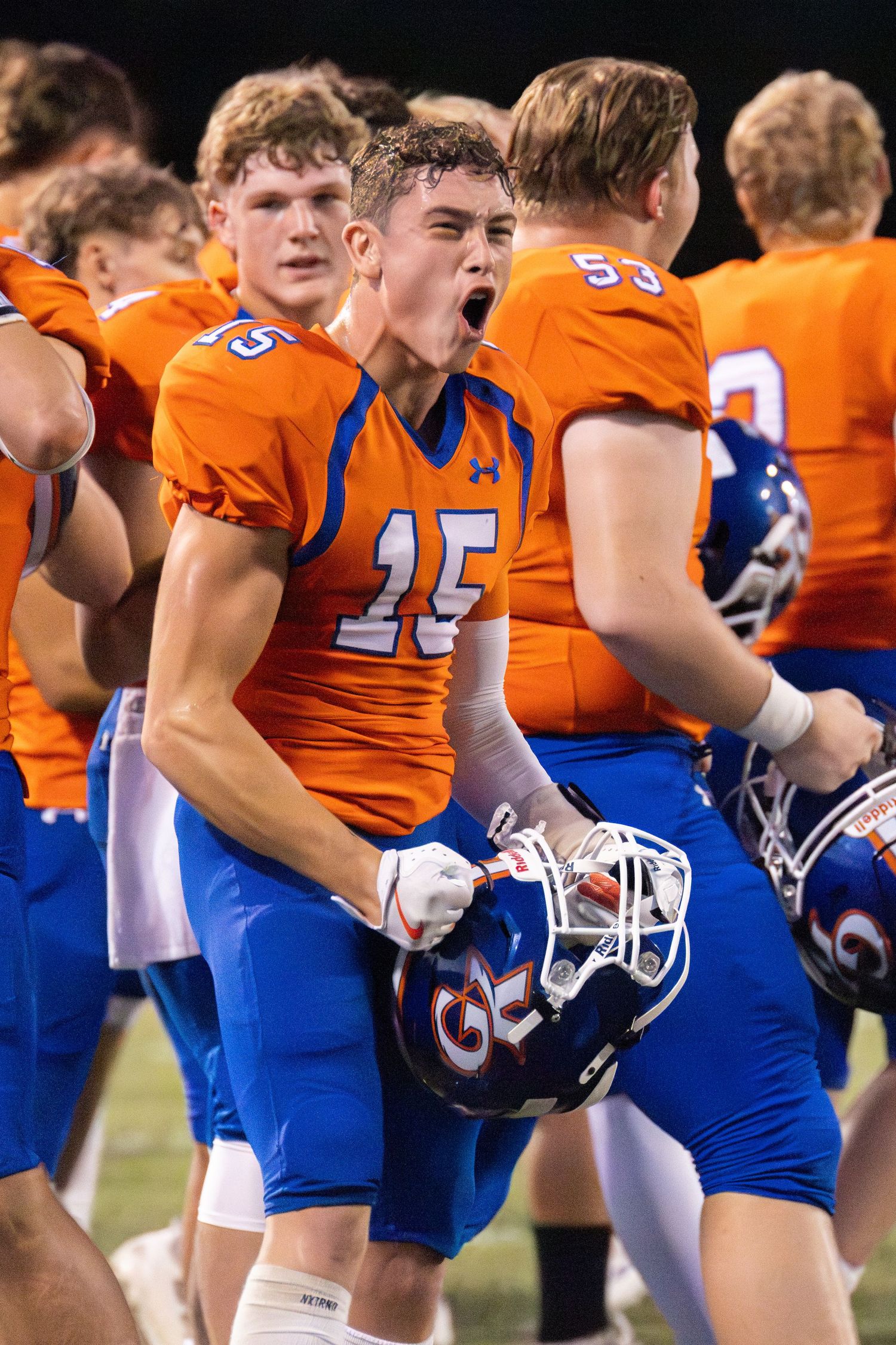 Football player #15 in orange and blue uniform celebrates victory, yelling with helmet in hand.