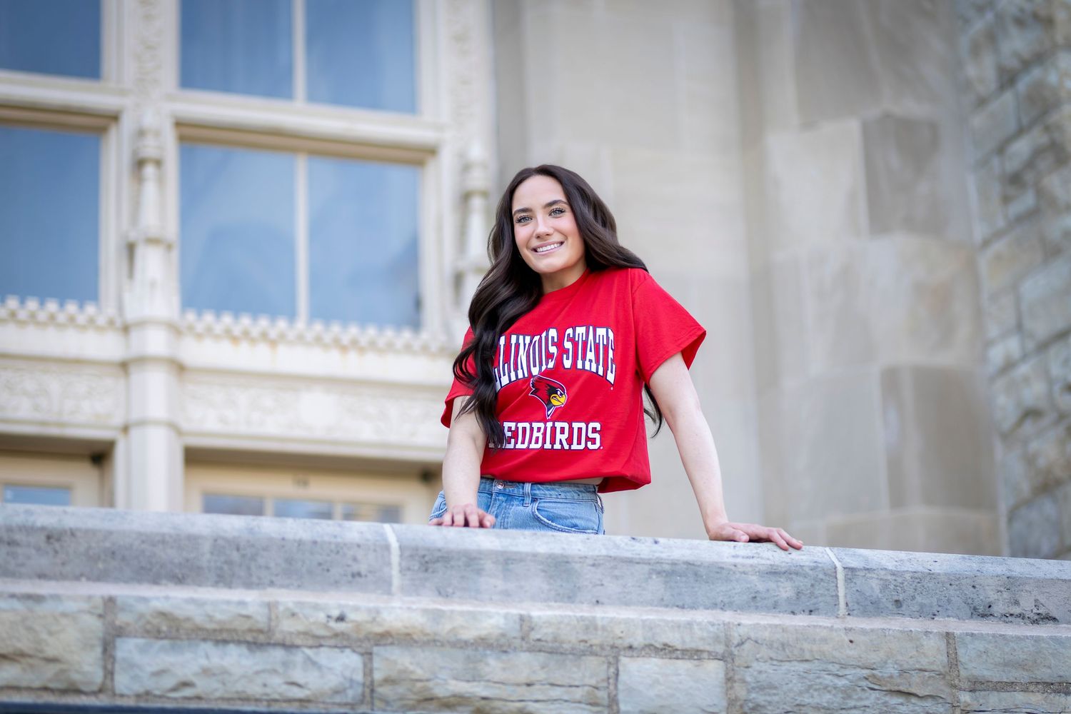 Young woman smiling in Illinois State Redbirds red t-shirt, posing on stone steps outside a campus building.
