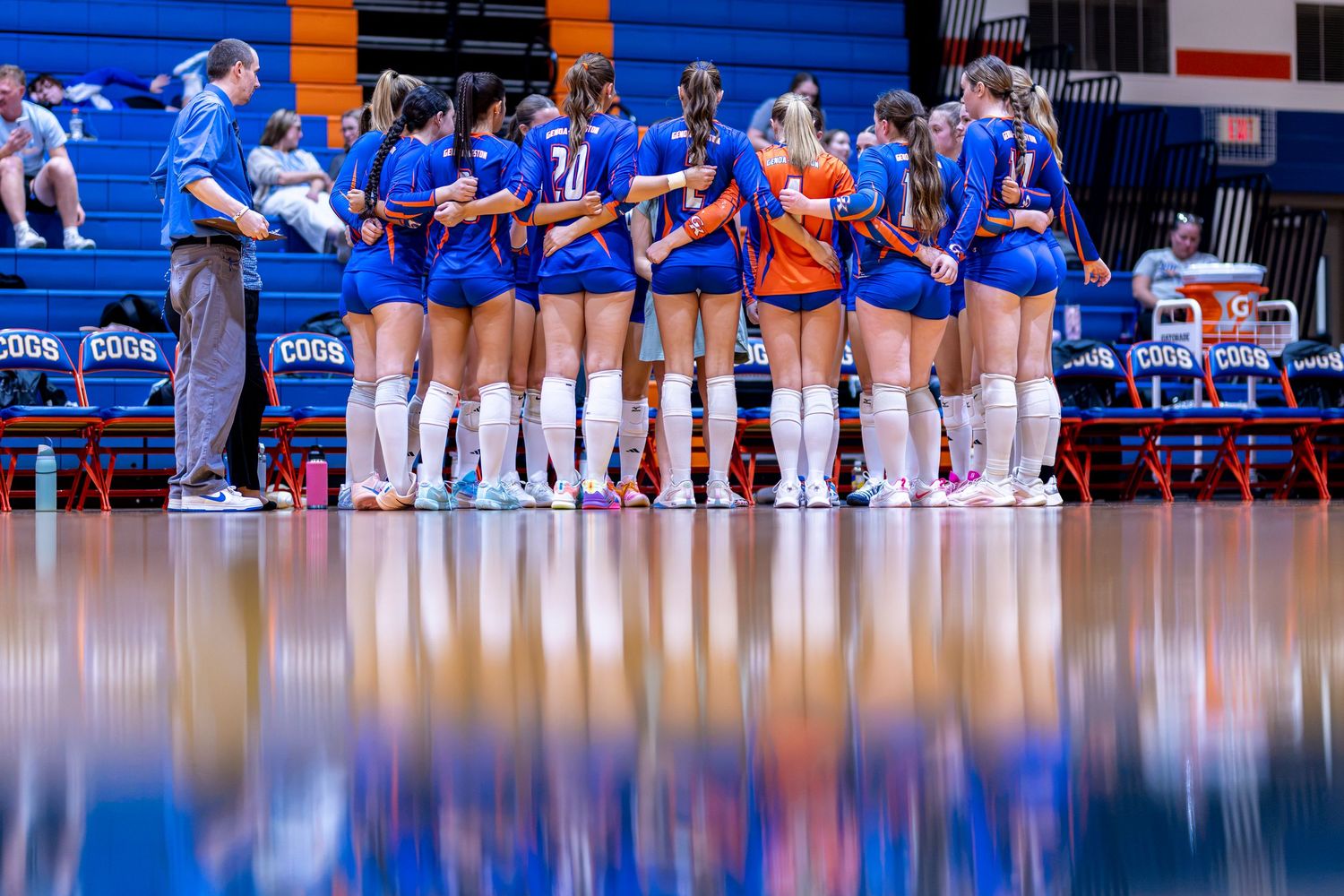 Boise State volleyball team huddles on court in blue uniforms, reflecting on polished hardwood floor during game.