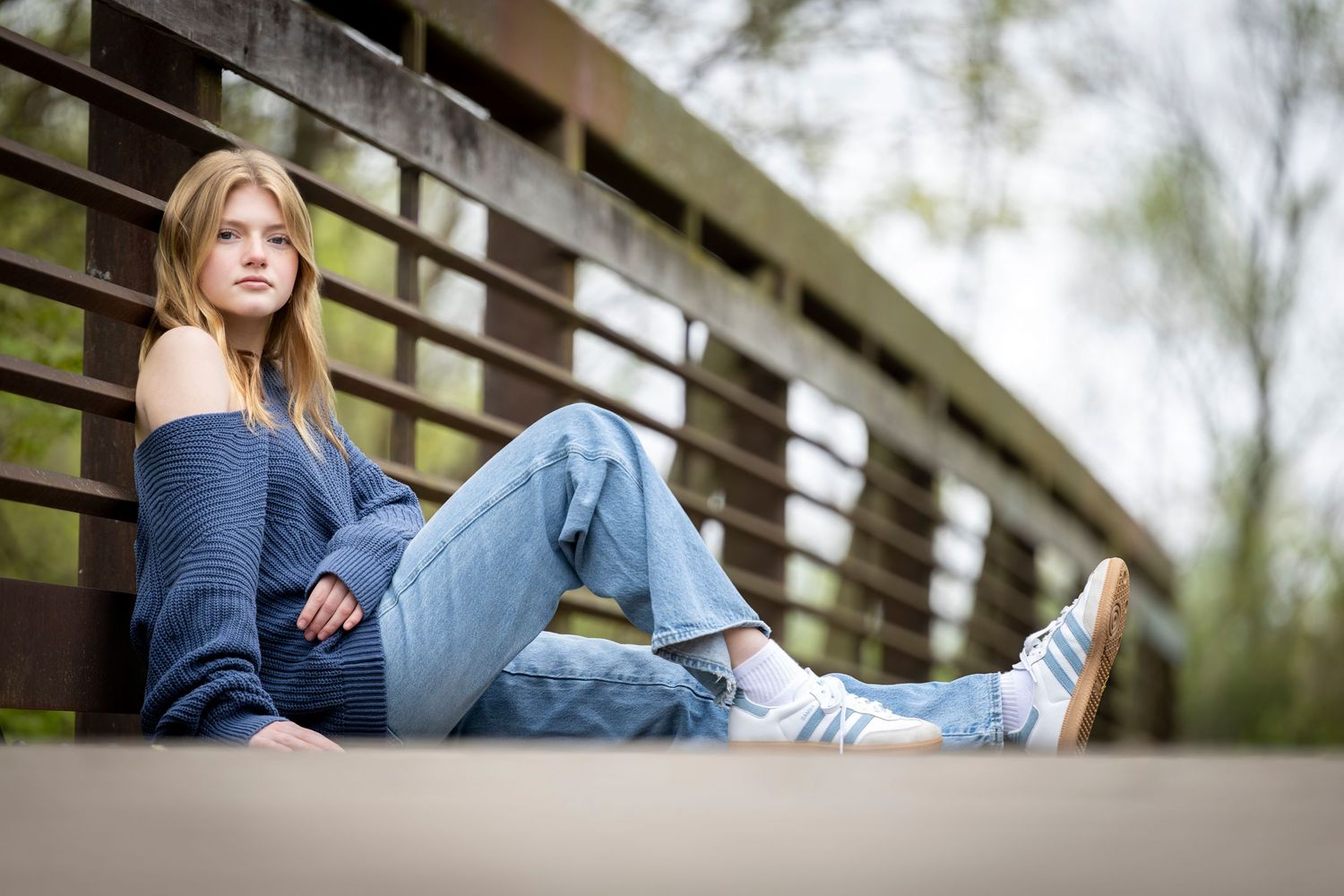 Young woman in blue sweater and jeans sitting relaxed on a wooden bridge outdoors.