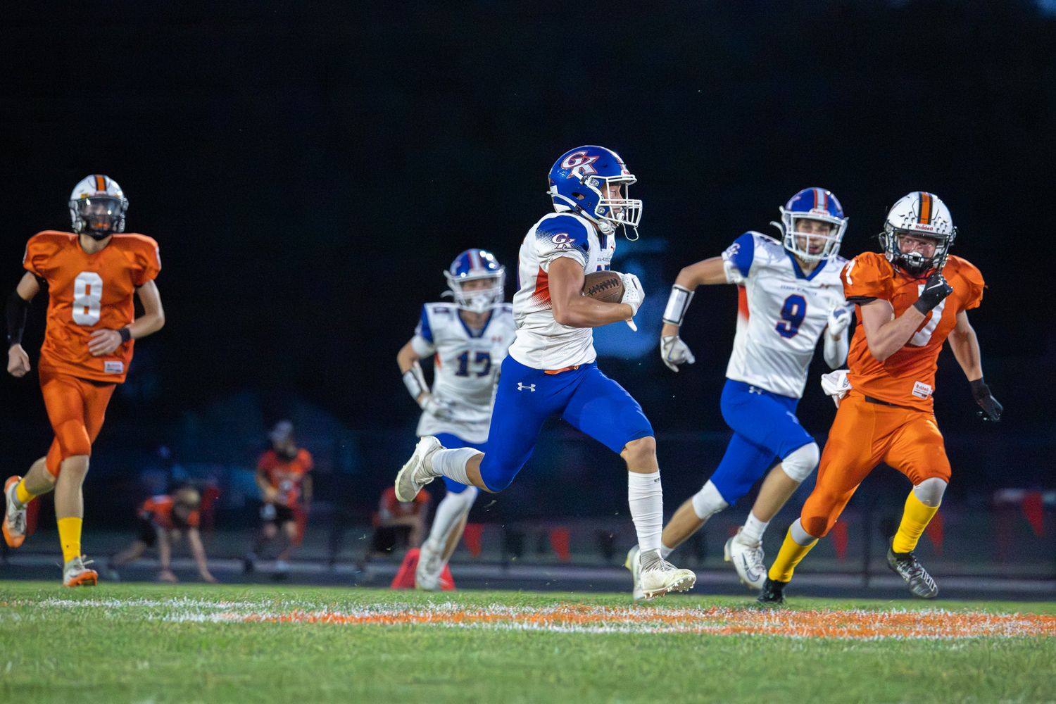 Football players in blue and orange jerseys line up in formation during a nighttime game.