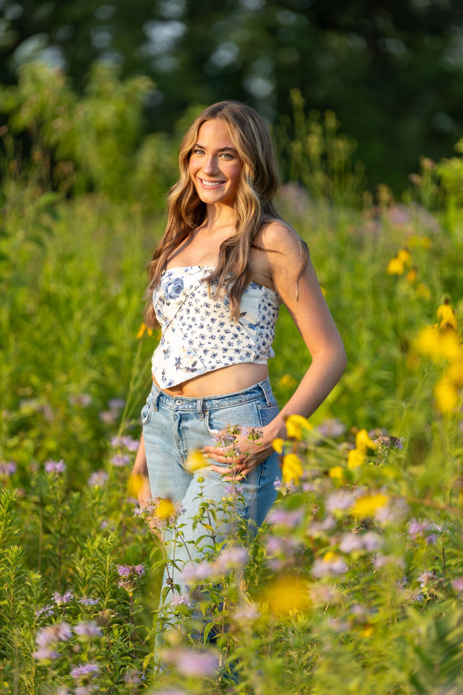 Young woman in floral crop top and jeans smiling in a sunny wildflower meadow with yellow and purple blooms.