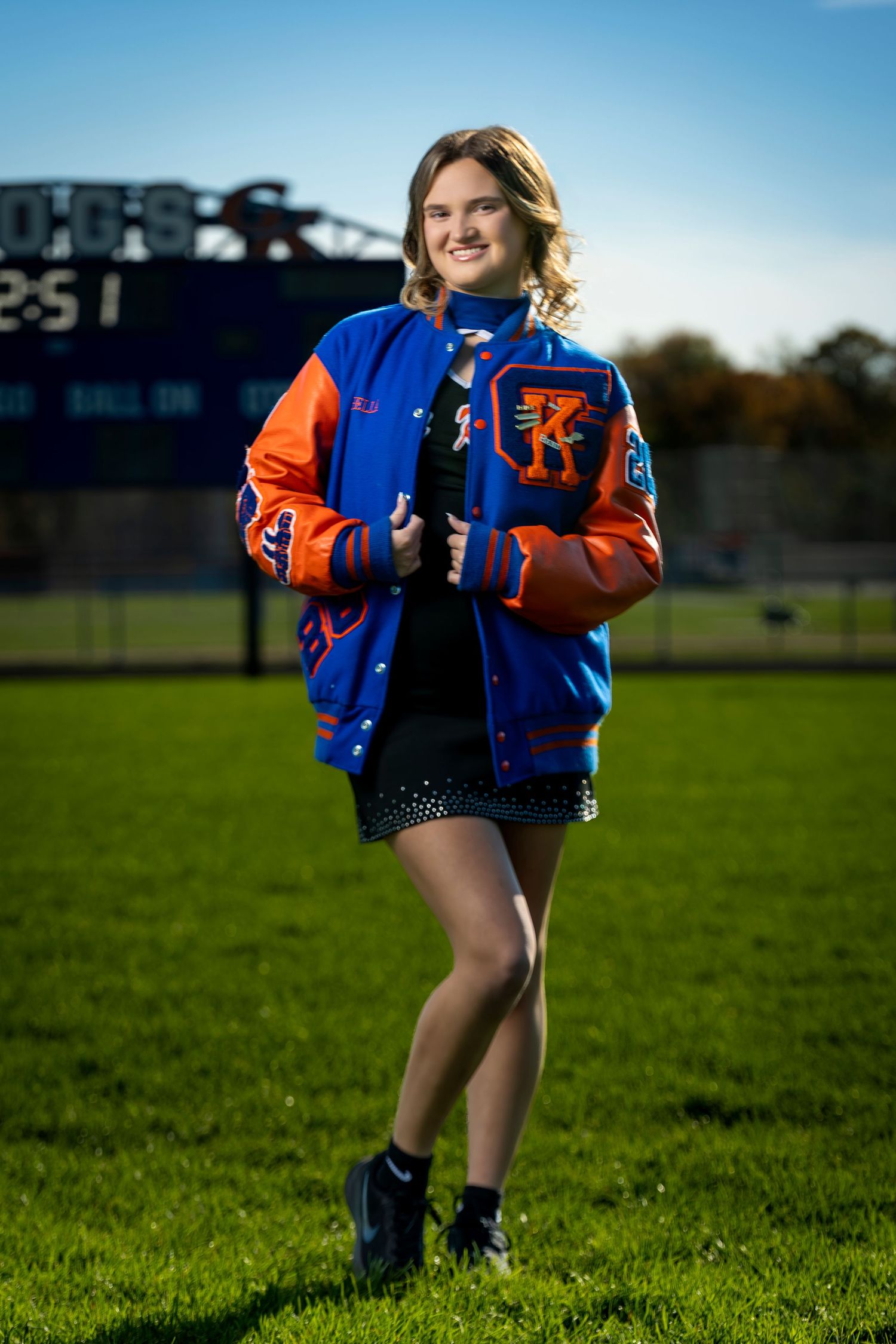Smiling teenage girl wearing a blue and orange varsity letterman jacket stands on a green football field.