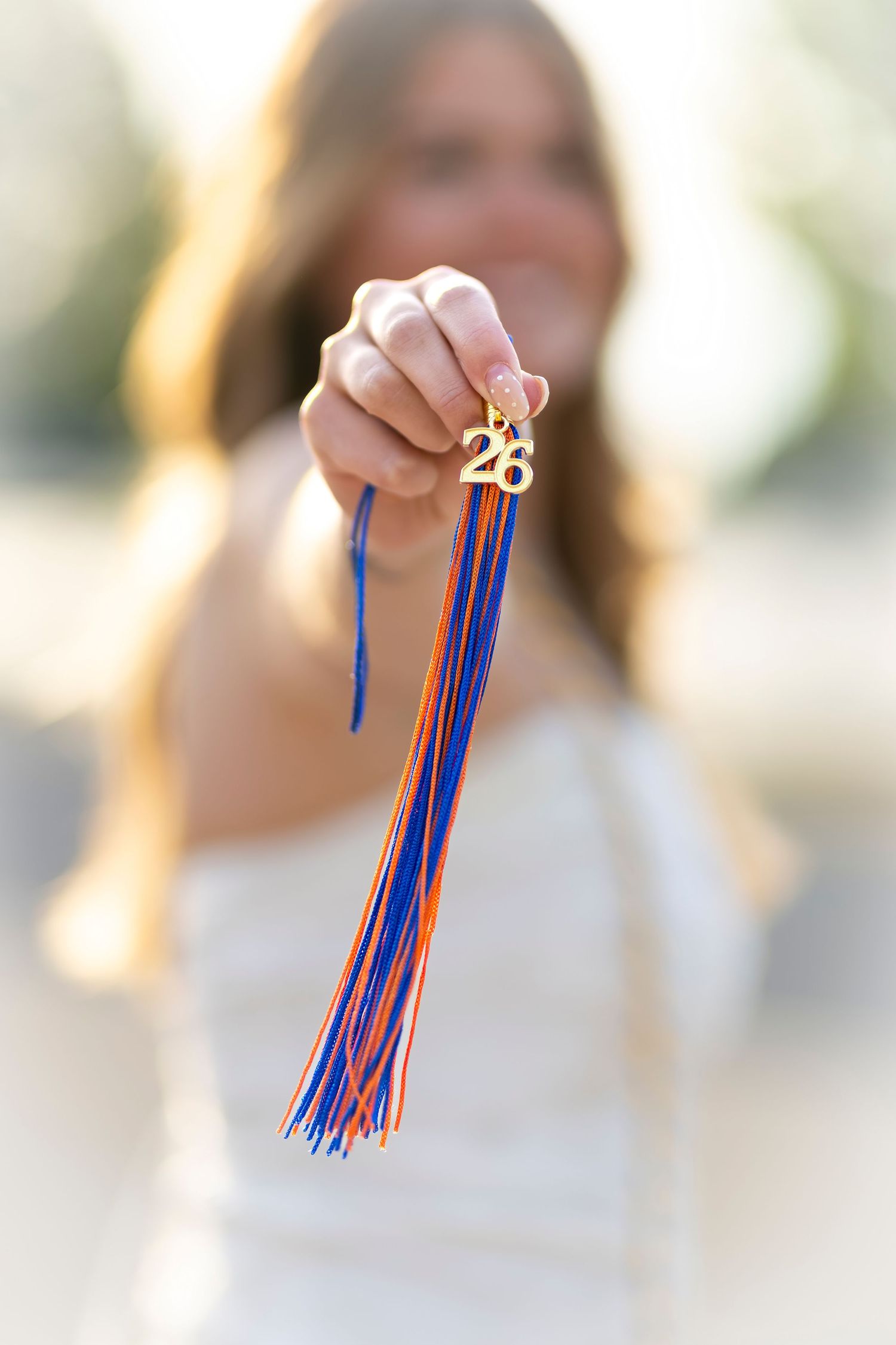 A young woman holds up a blue and orange graduation tassel with a gold '26' charm for her senior photos.
