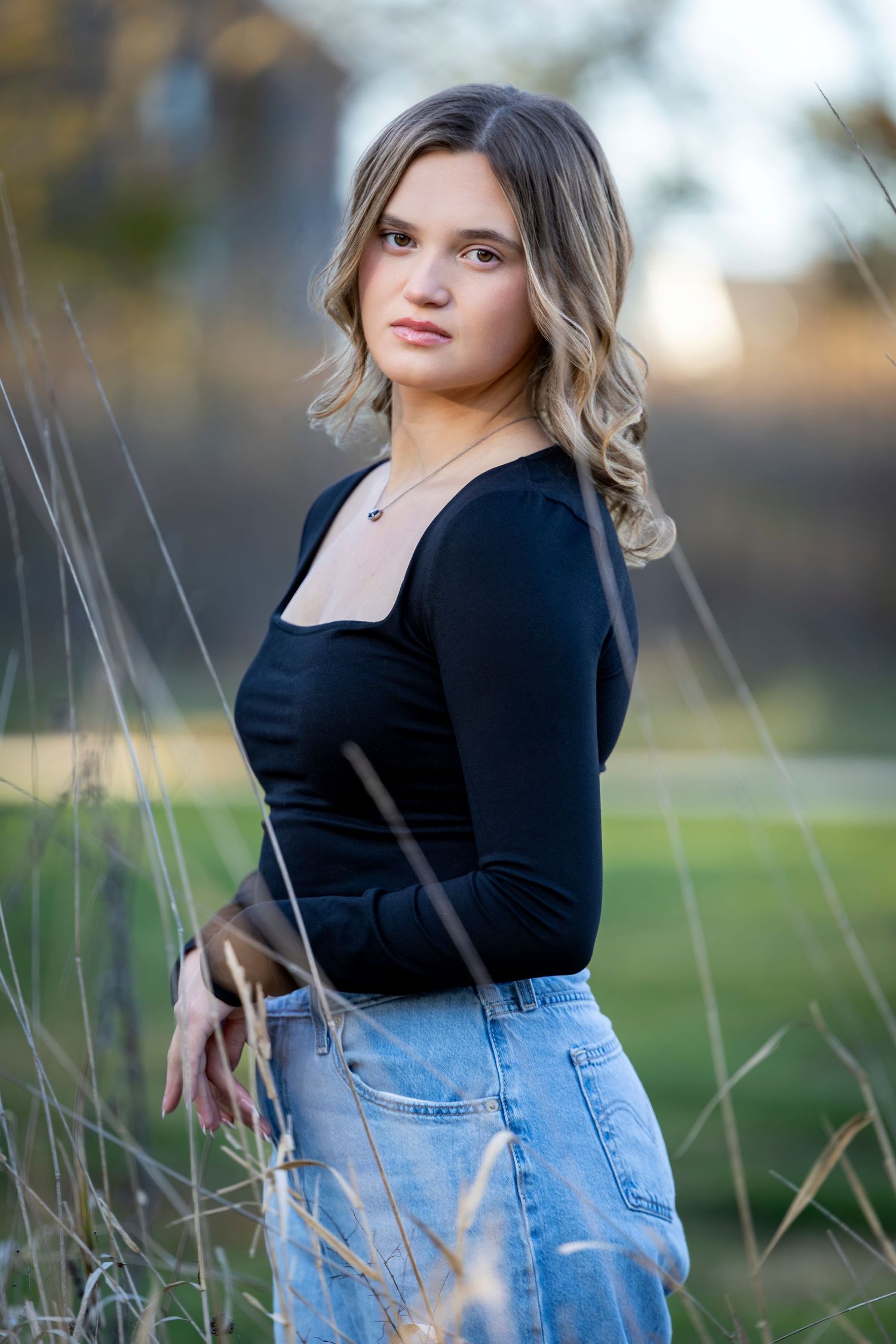 Young woman in black top and jeans poses outdoors among tall grass in warm autumn light.
