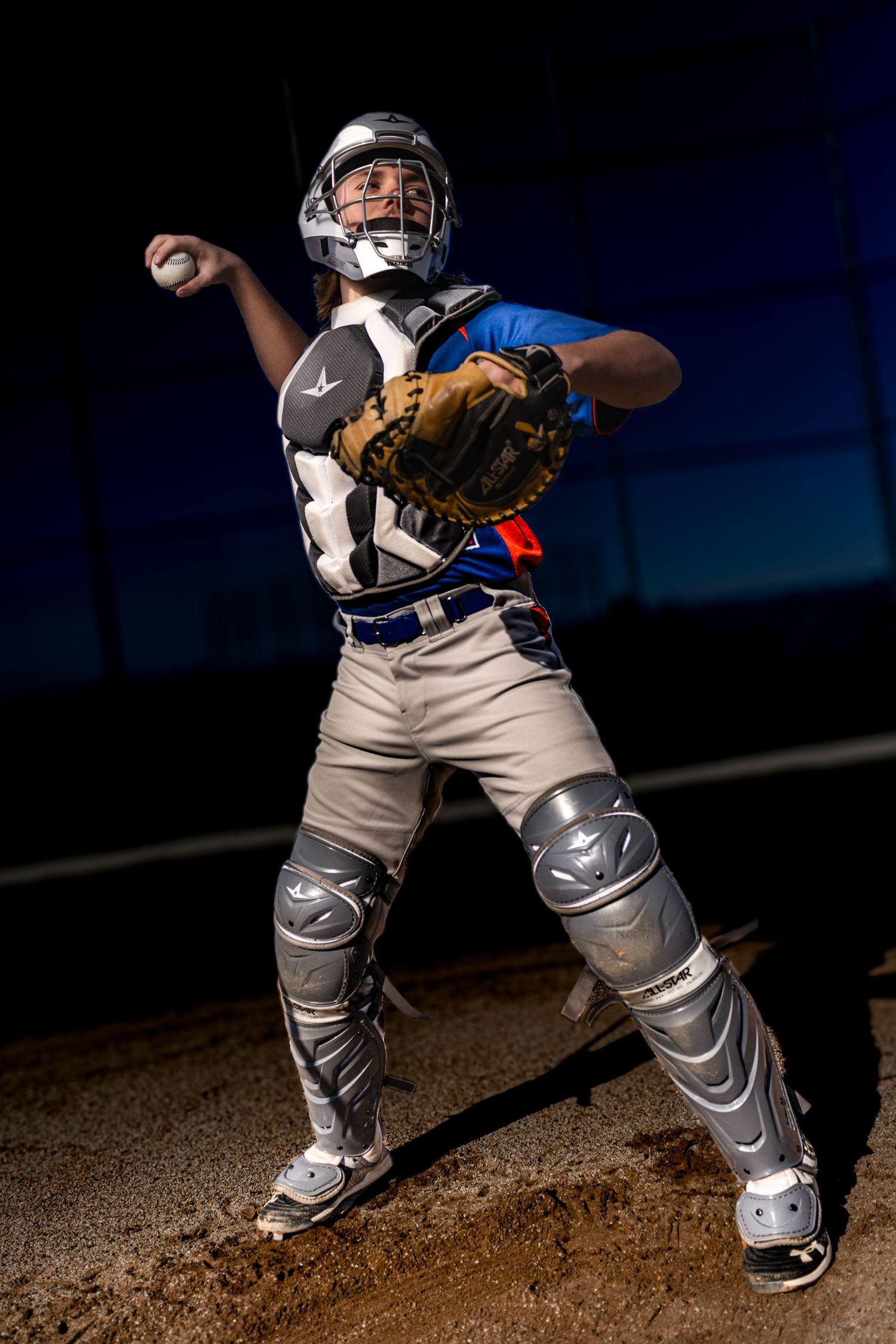 Baseball catcher in full gear throwing ball on field, wearing helmet, chest protector, and shin guards.