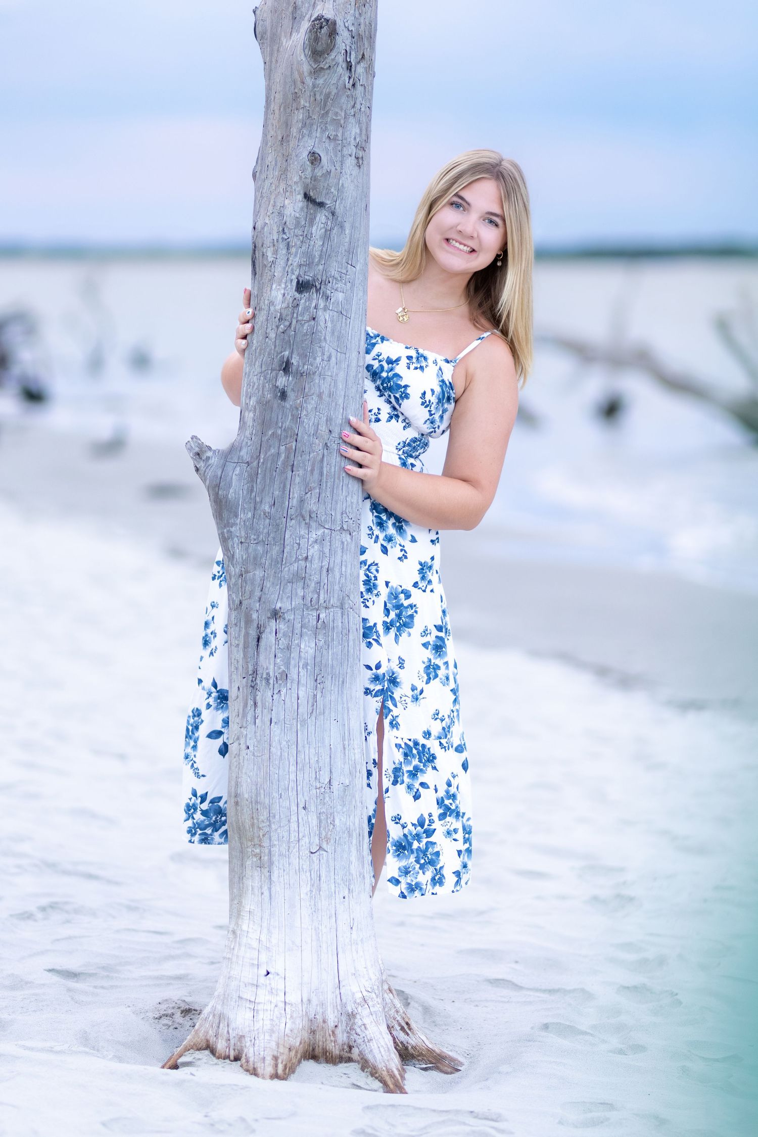 Smiling blonde woman in blue floral sundress posing by driftwood on white sandy beach.