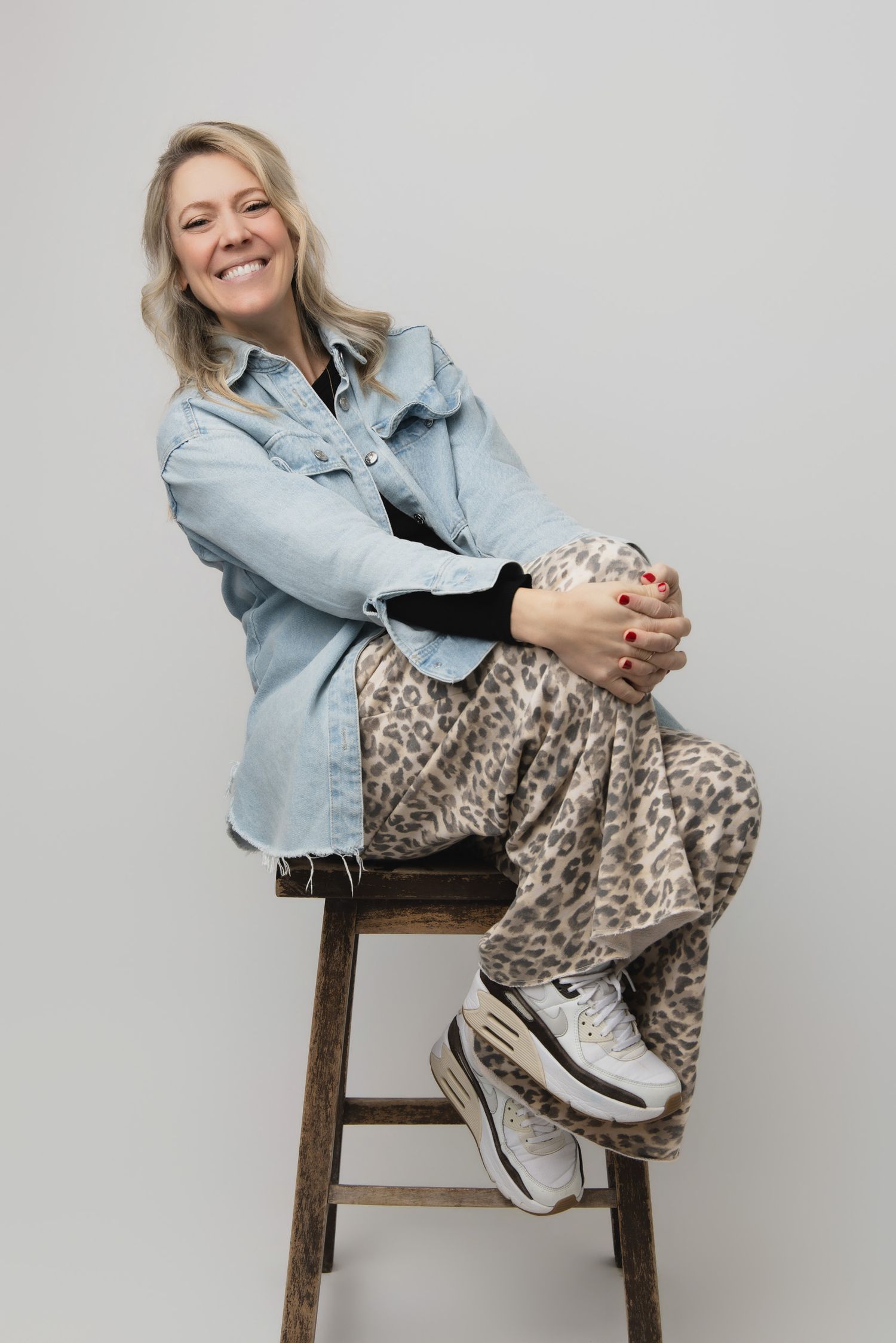 A person in a denim jacket and leopard print pants sits casually on a wooden stool against a gray background.