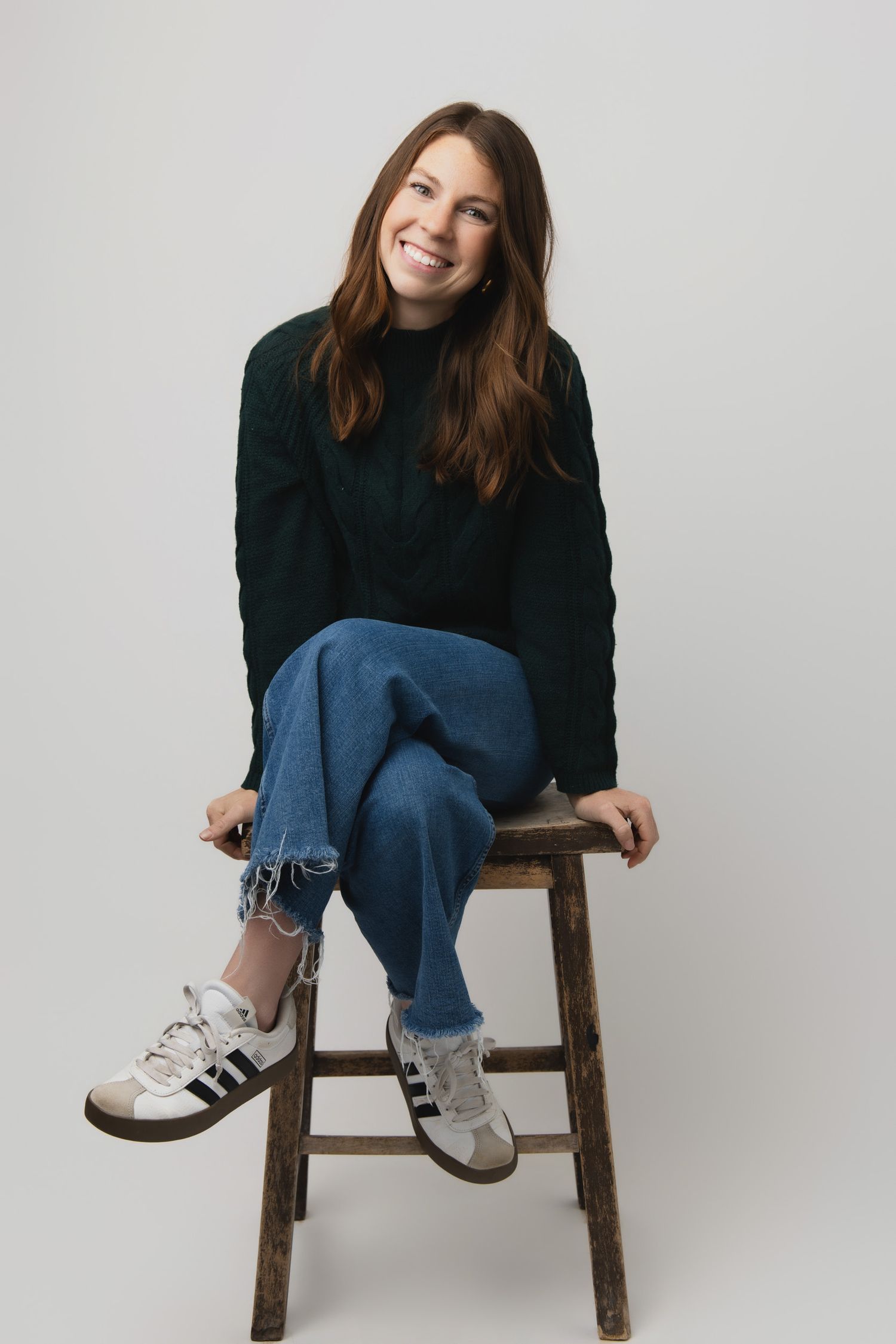Casual portrait on wooden stool wearing dark sweater, distressed jeans and white sneakers against light gray background.
