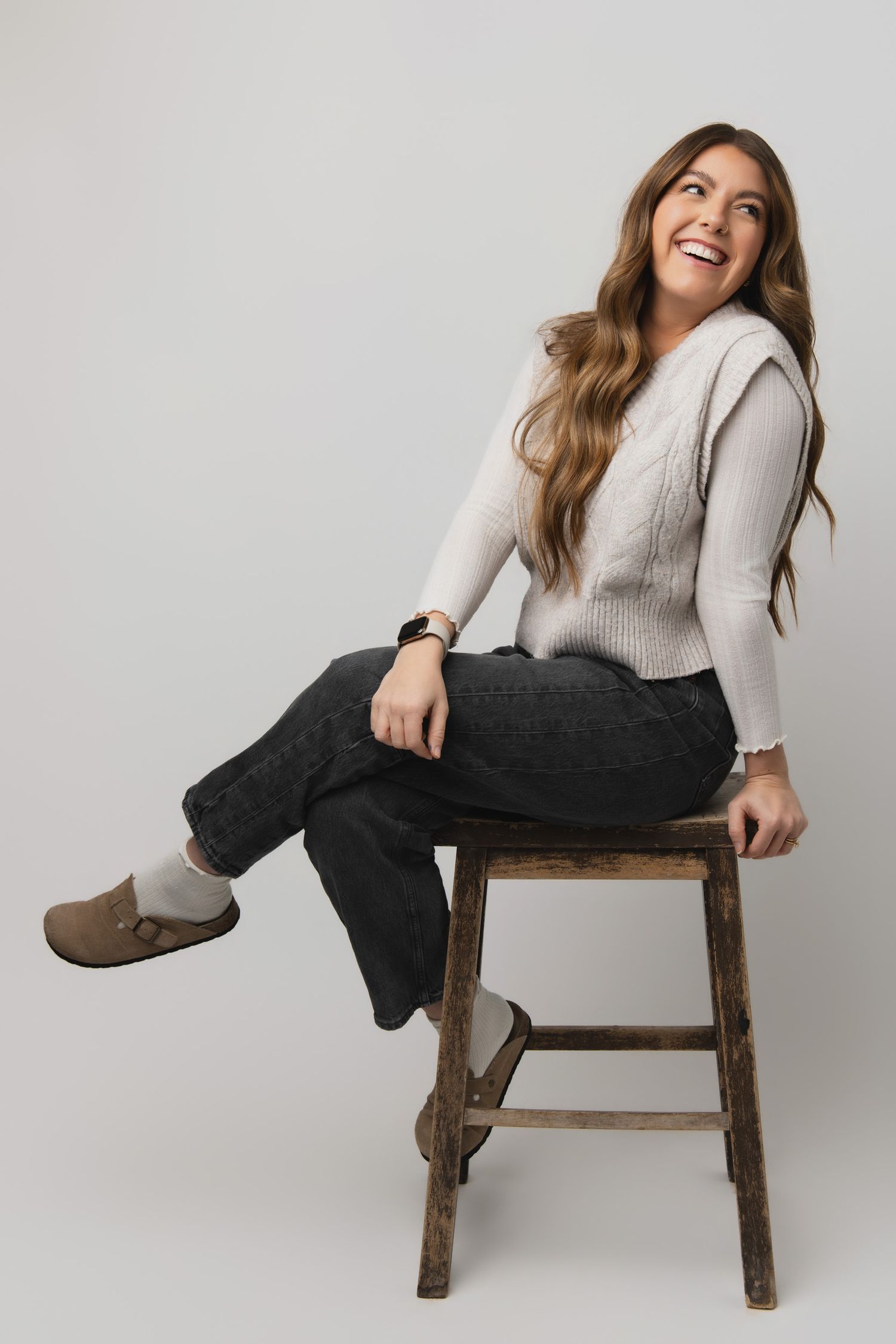 Casual portrait of someone sitting on wooden stool wearing cream sweater, dark pants and brown shoes against gray background.