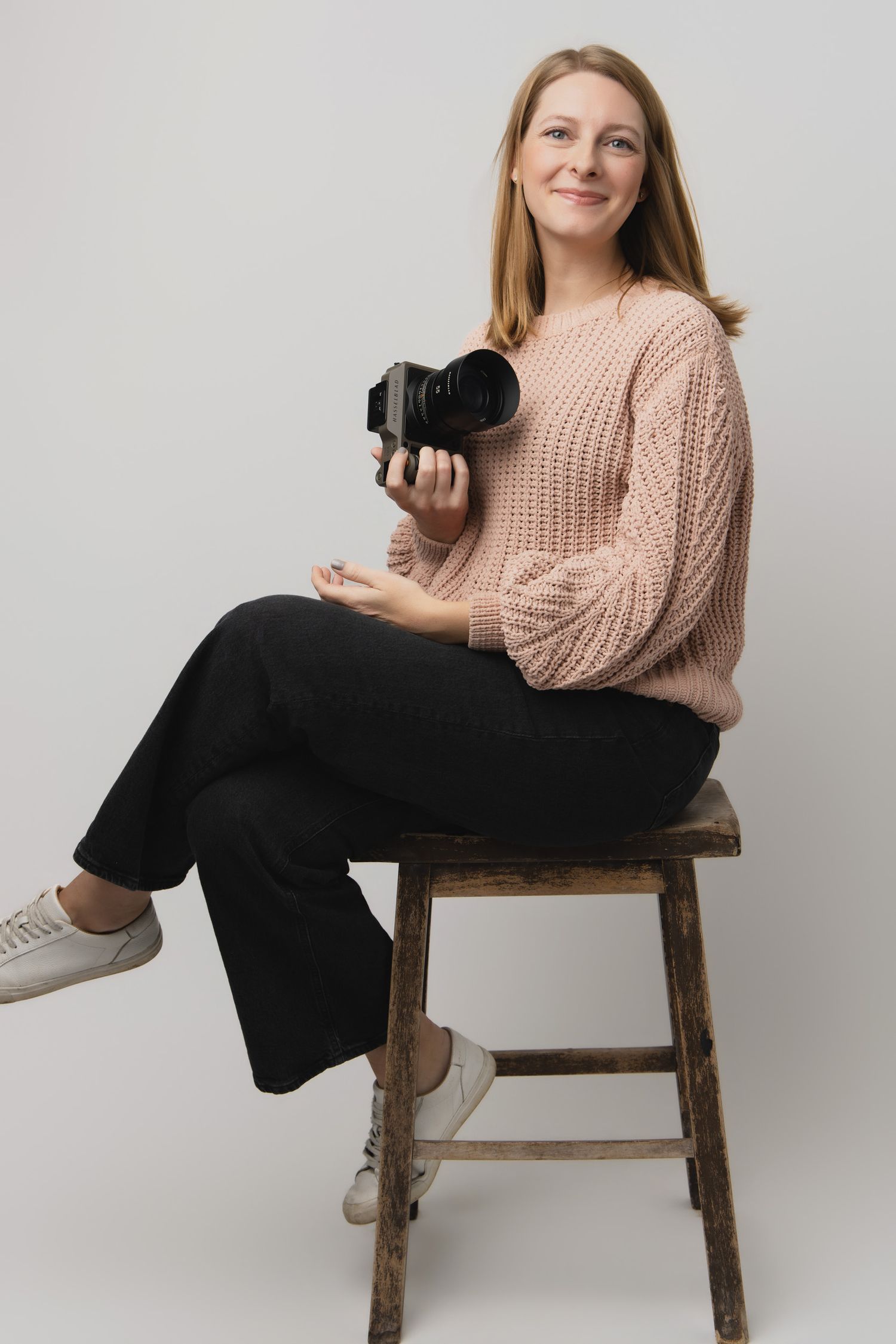 A photographer in a pink knit sweater and black pants poses casually on a wooden stool against a white backdrop.