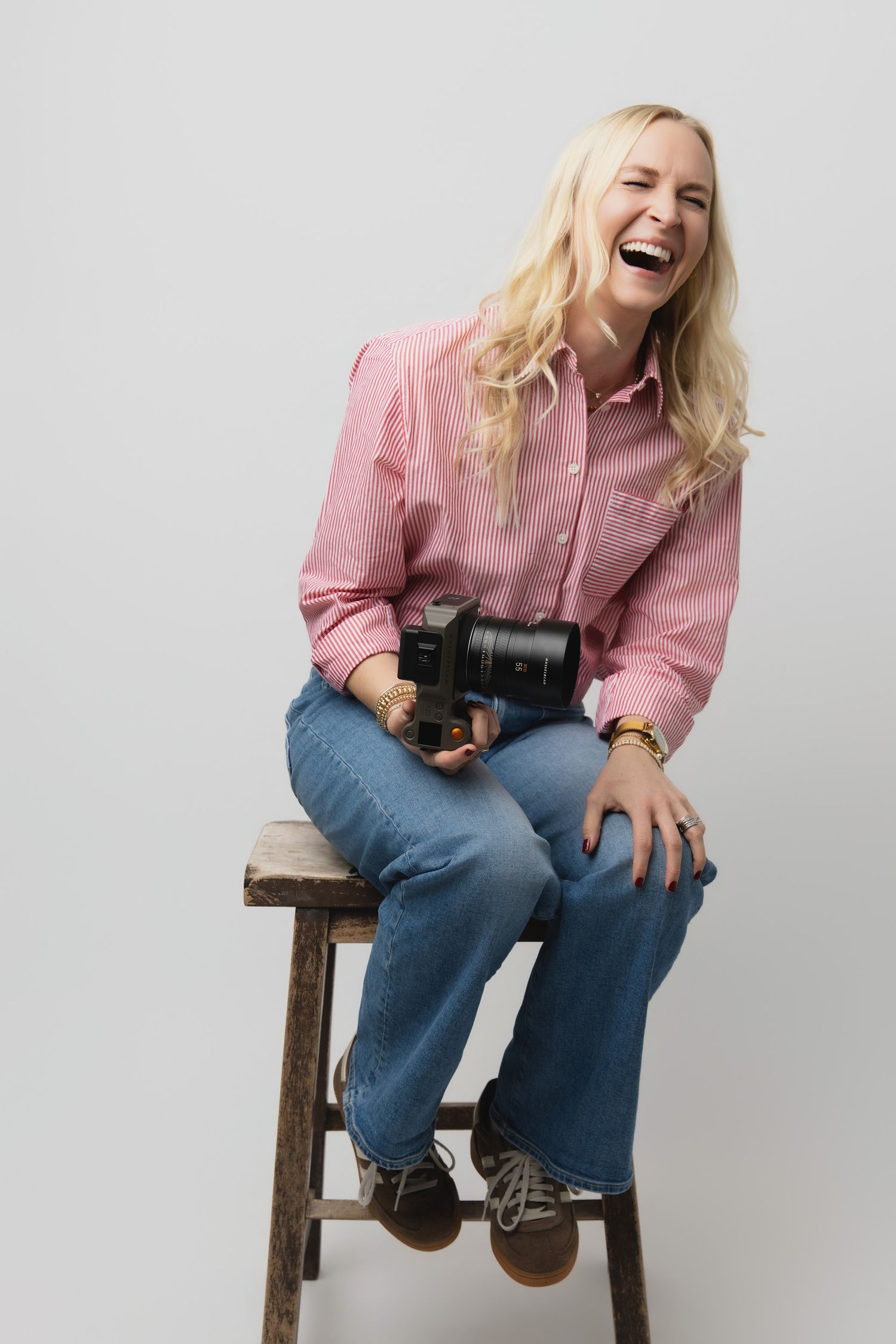 Person wearing pink shirt and jeans sits on wooden stool laughing joyfully against white background.
