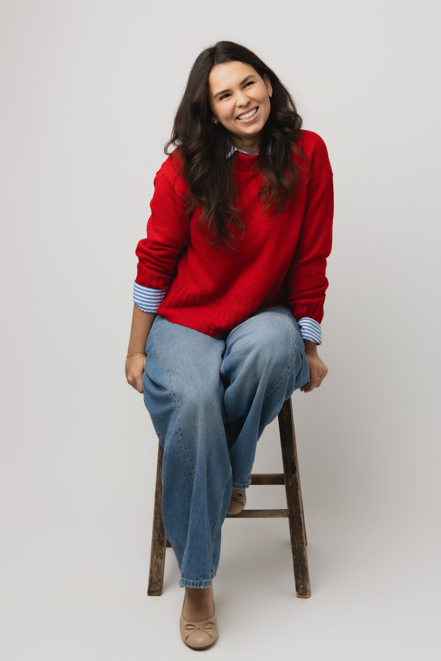 Person wearing a cozy red sweater and relaxed jeans sits casually on a wooden stool against white background.