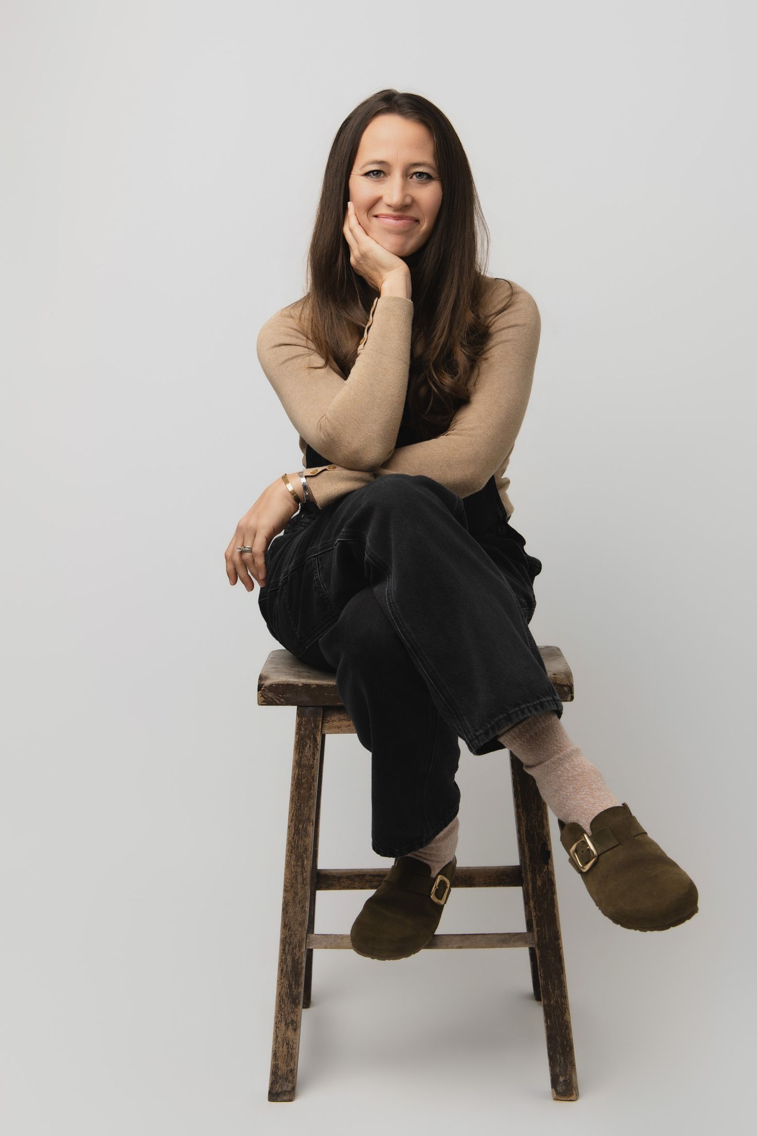 Portrait of someone seated casually on a wooden stool wearing a beige sweater, black pants and brown shoes against white backdrop.