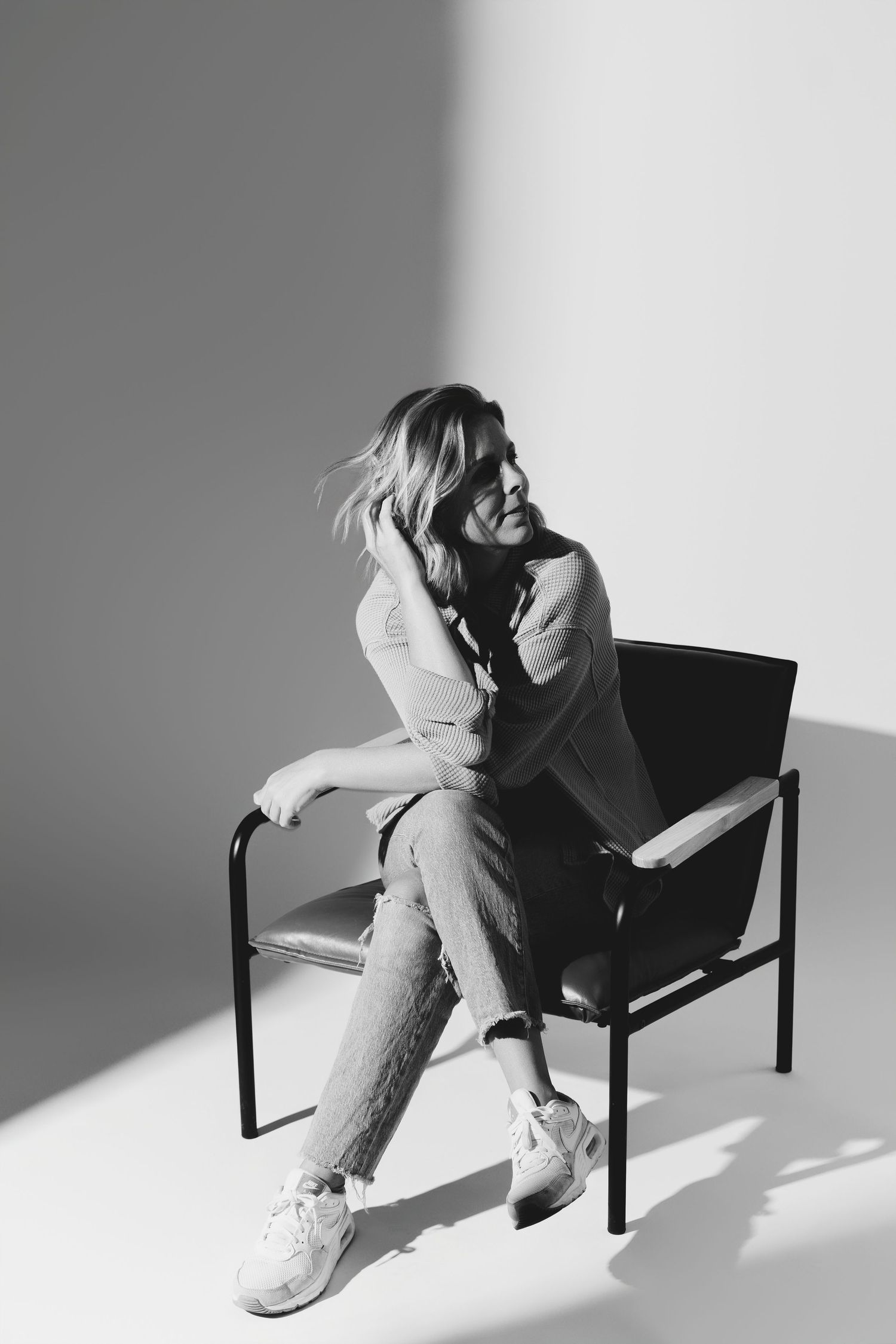 A black and white portrait of someone seated casually in a chair wearing jeans and sneakers while laughing.