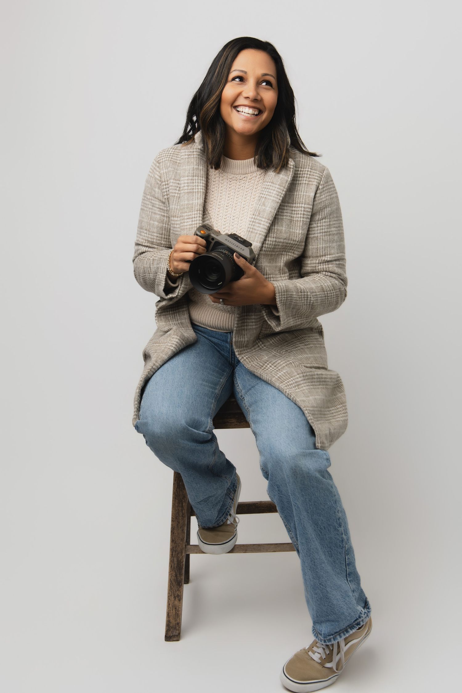 Professional photographer wearing plaid blazer and jeans sits casually with camera on wooden stool against white backdrop.