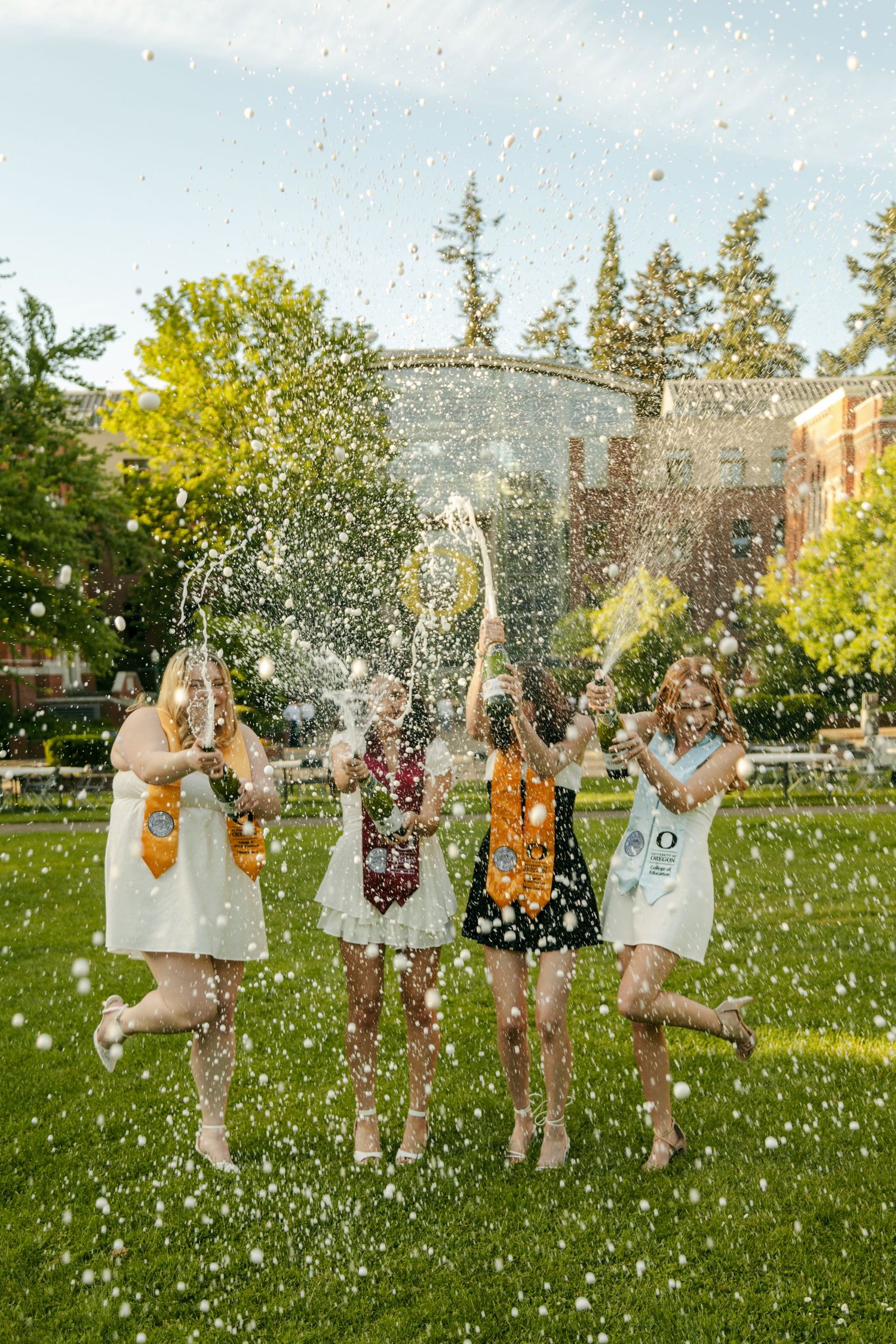 Four college graduates in white dresses celebrate outdoors with confetti and champagne on a sunny campus lawn.