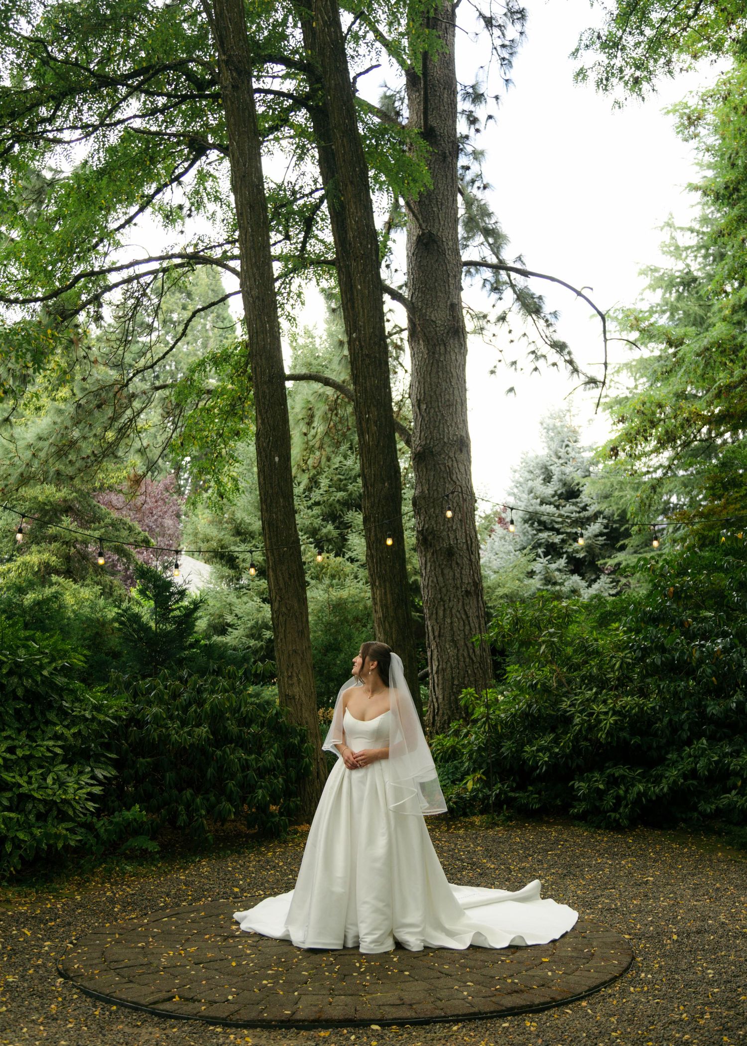 Bride in white gown with train stands in lush garden surrounded by tall trees on a cloudy day.