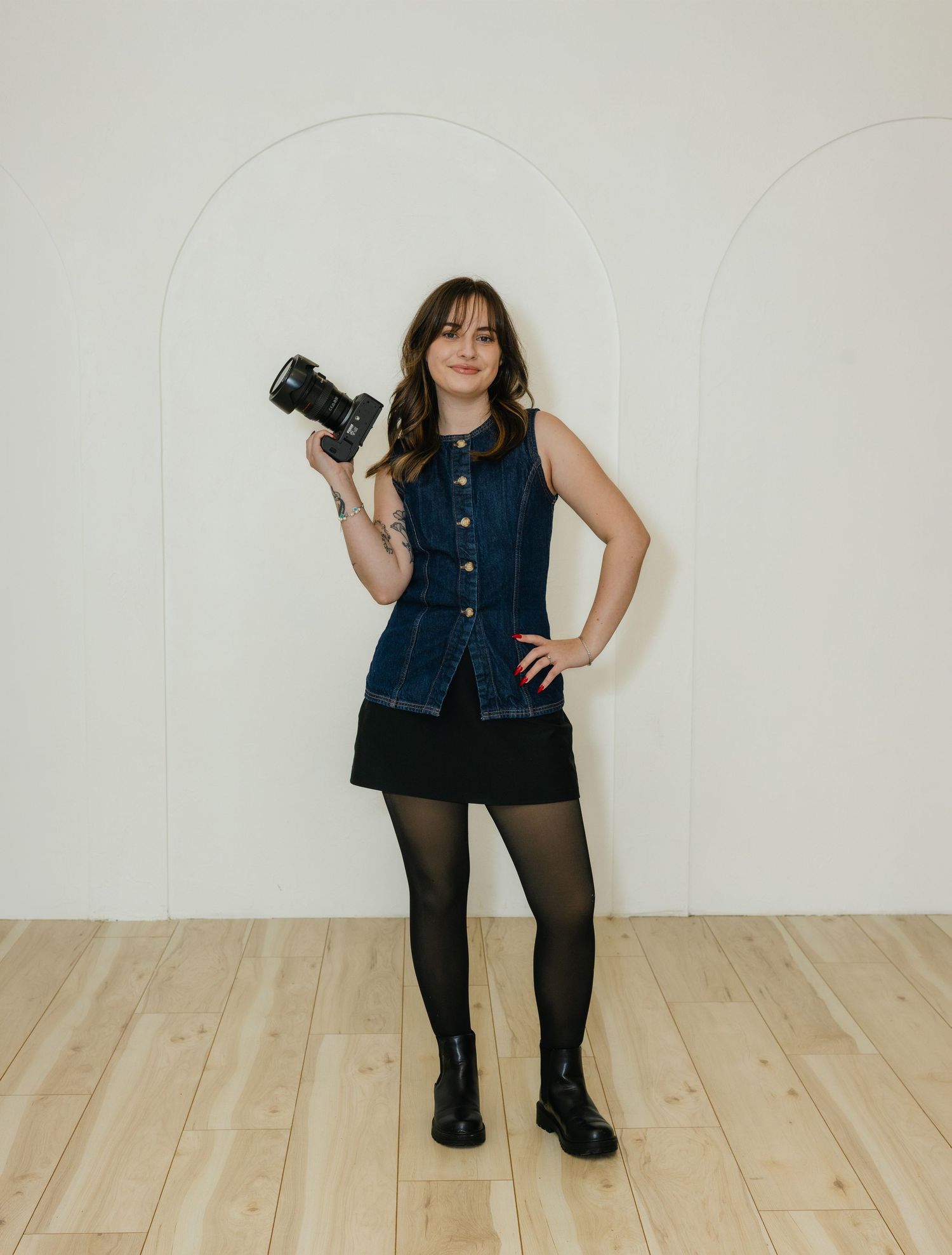A photographer in a denim vest, black skirt and tights stands against a white wall while holding a professional camera.