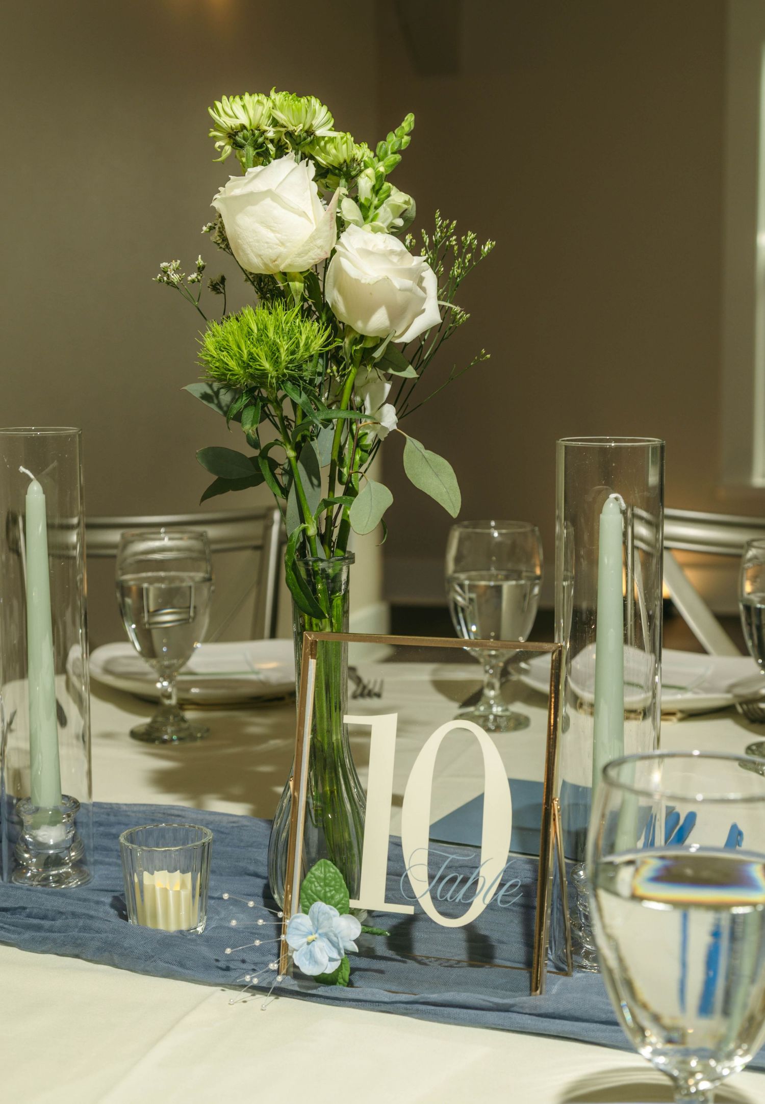 Elegant wedding table centerpiece with white roses, green flowers in tall vase, table number 10, blue runner, candles.