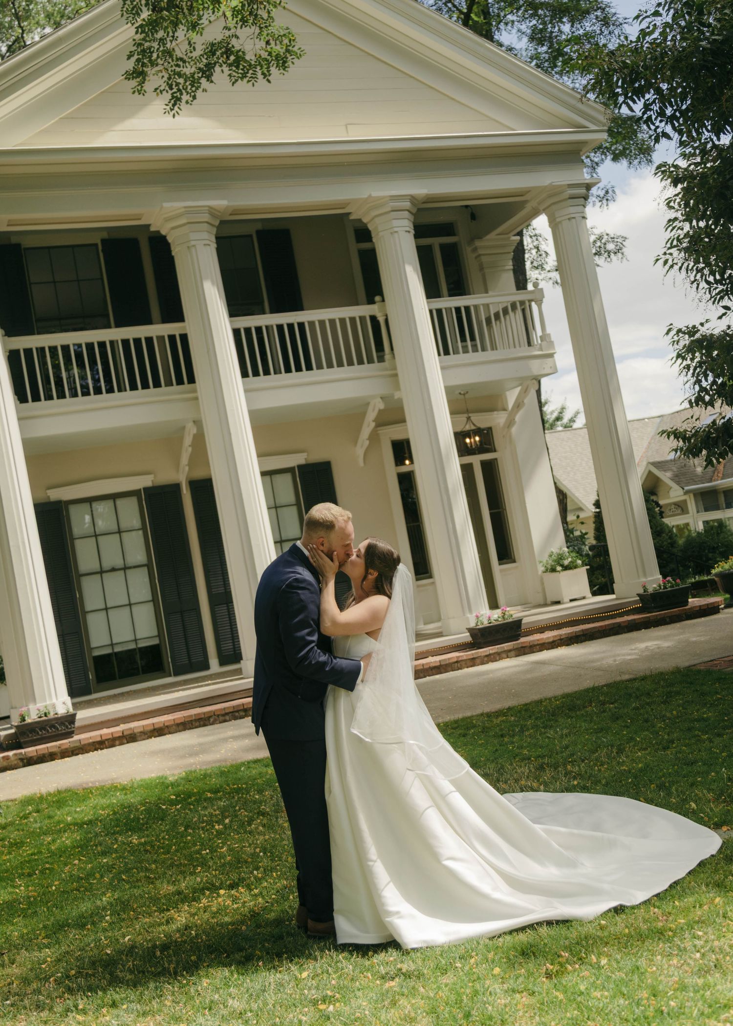 Bride and groom share a romantic kiss on the lawn of an elegant white colonial mansion with tall pillars on their wedding day.