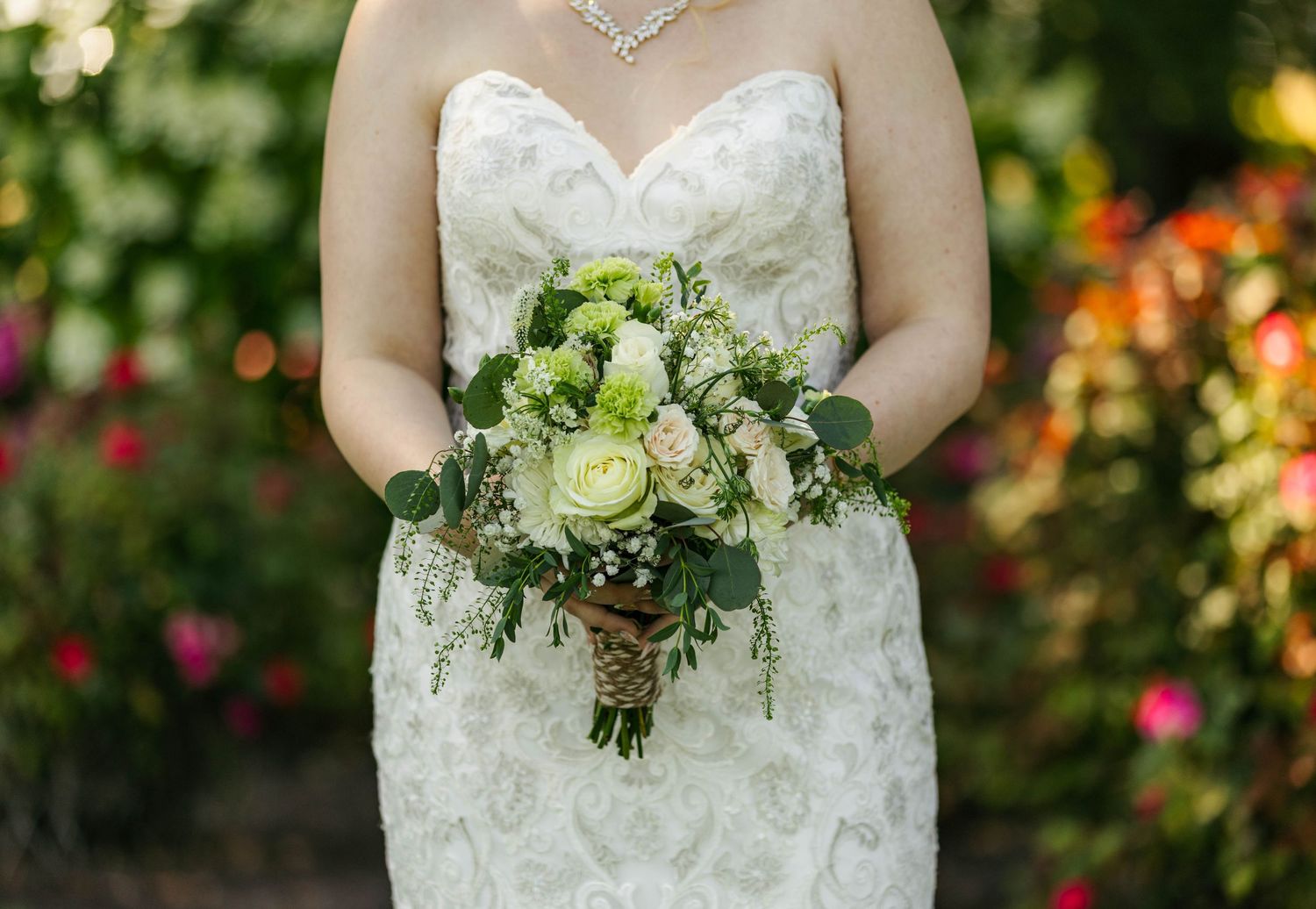 Bride in strapless lace wedding gown holding green and white rose bouquet with eucalyptus in garden setting.