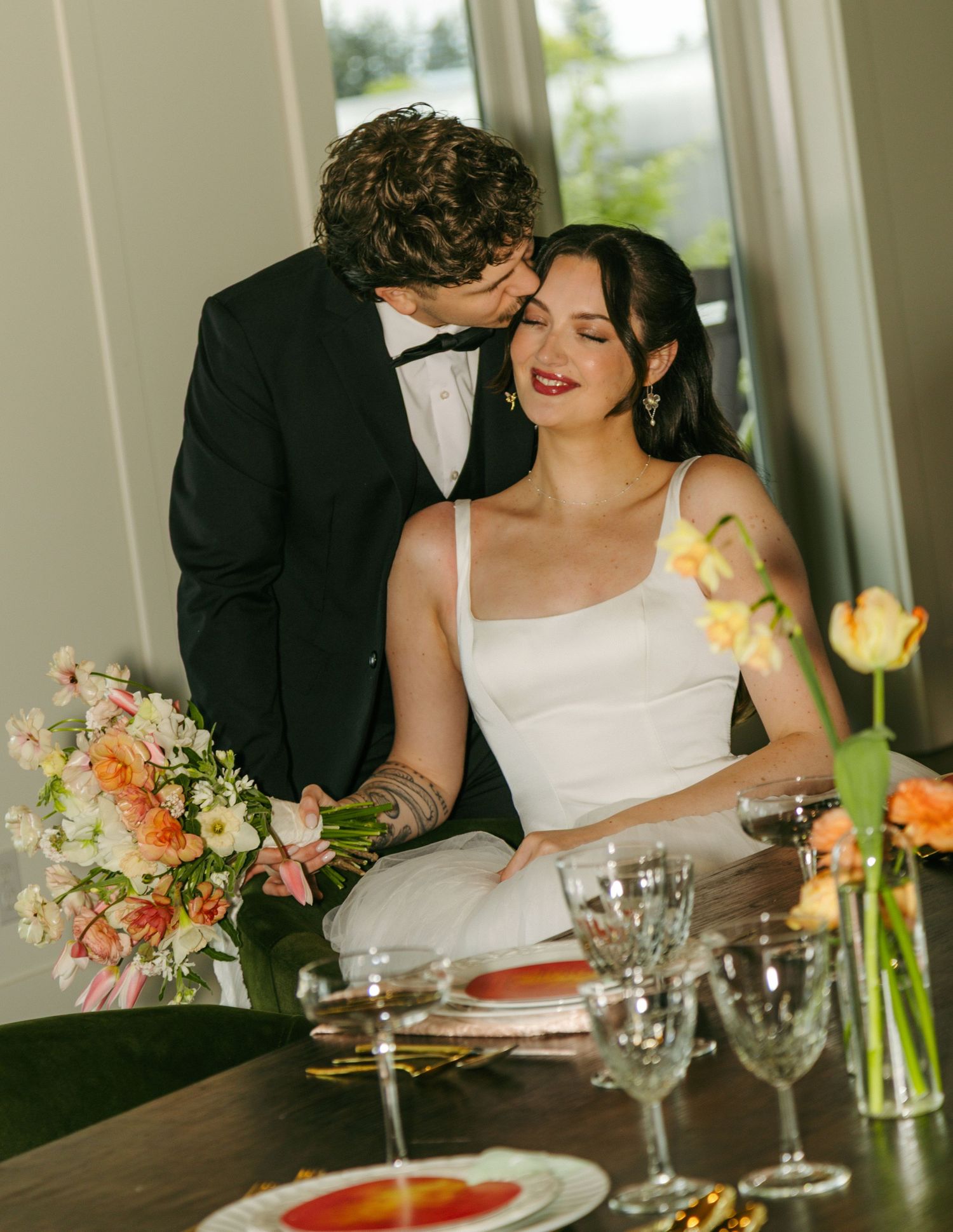 Bride in white gown sits at decorated table as groom in black tuxedo kisses her cheek at elegant wedding reception.
