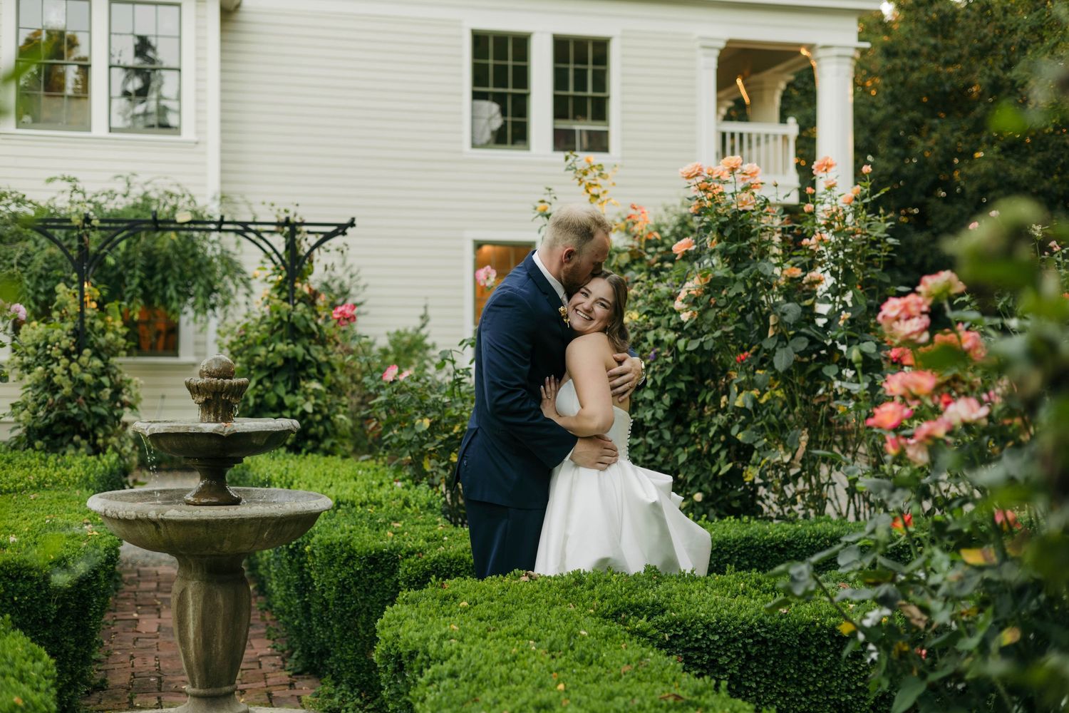 Bride and groom share a romantic kiss in a lush garden with blooming roses and a stone fountain near a white estate.