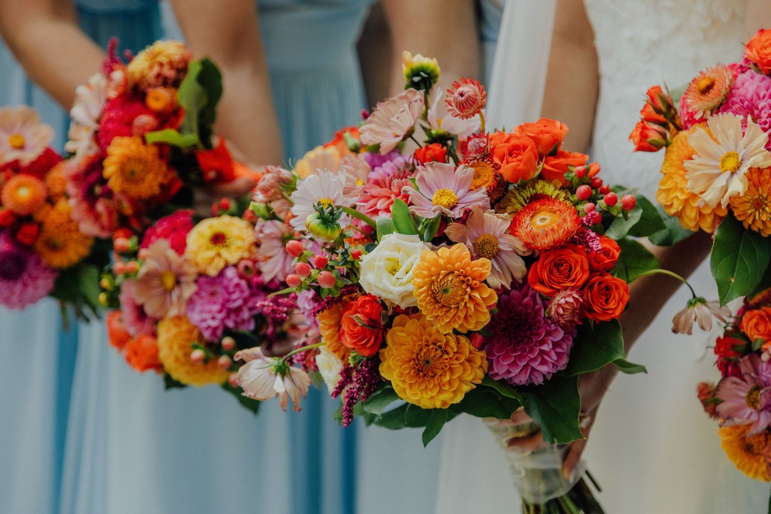 Vibrant bouquets of orange, pink and purple dahlias and mixed flowers held against light blue dresses at a wedding.