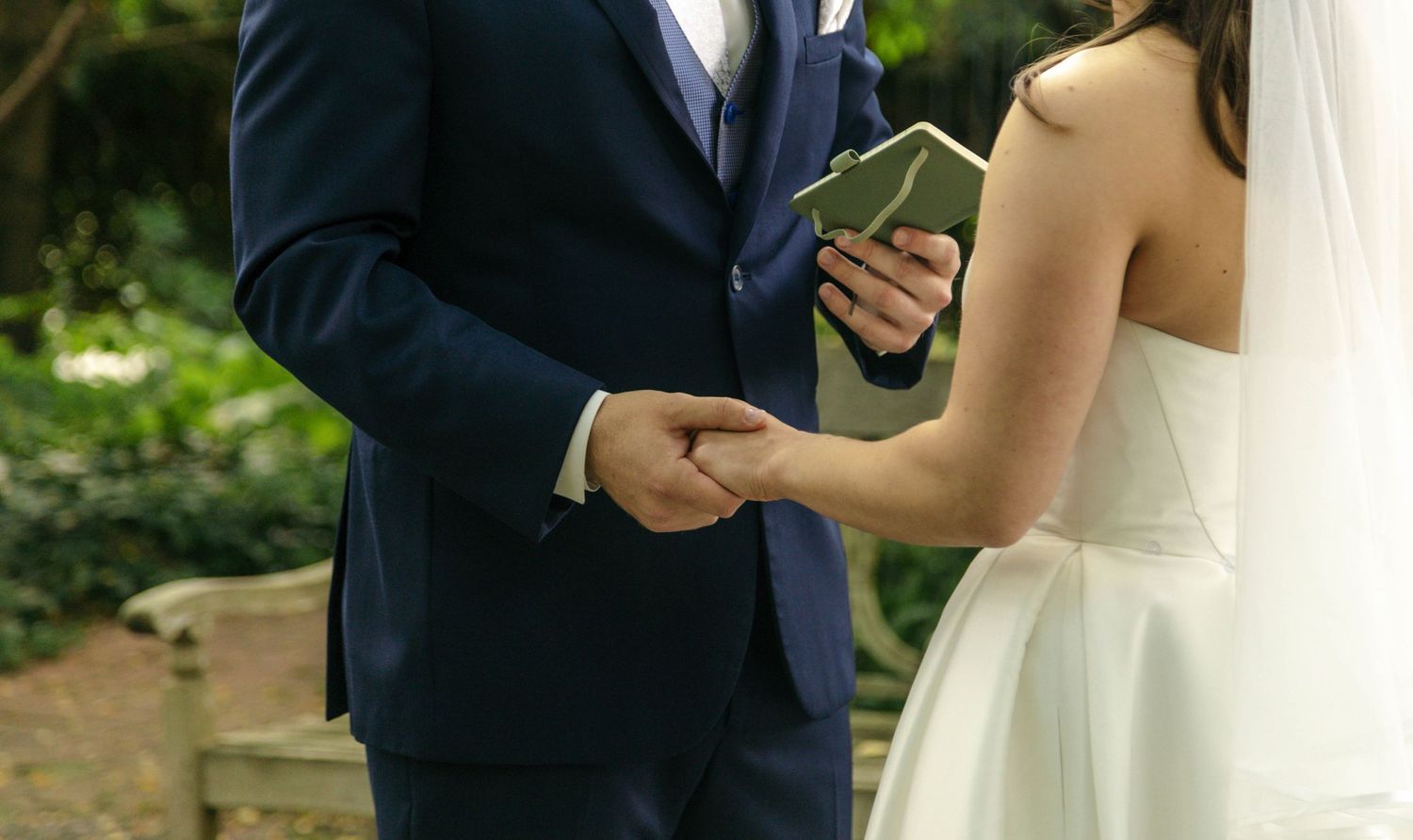 Groom in navy suit holding bride's hand during outdoor wedding ceremony vow exchange.