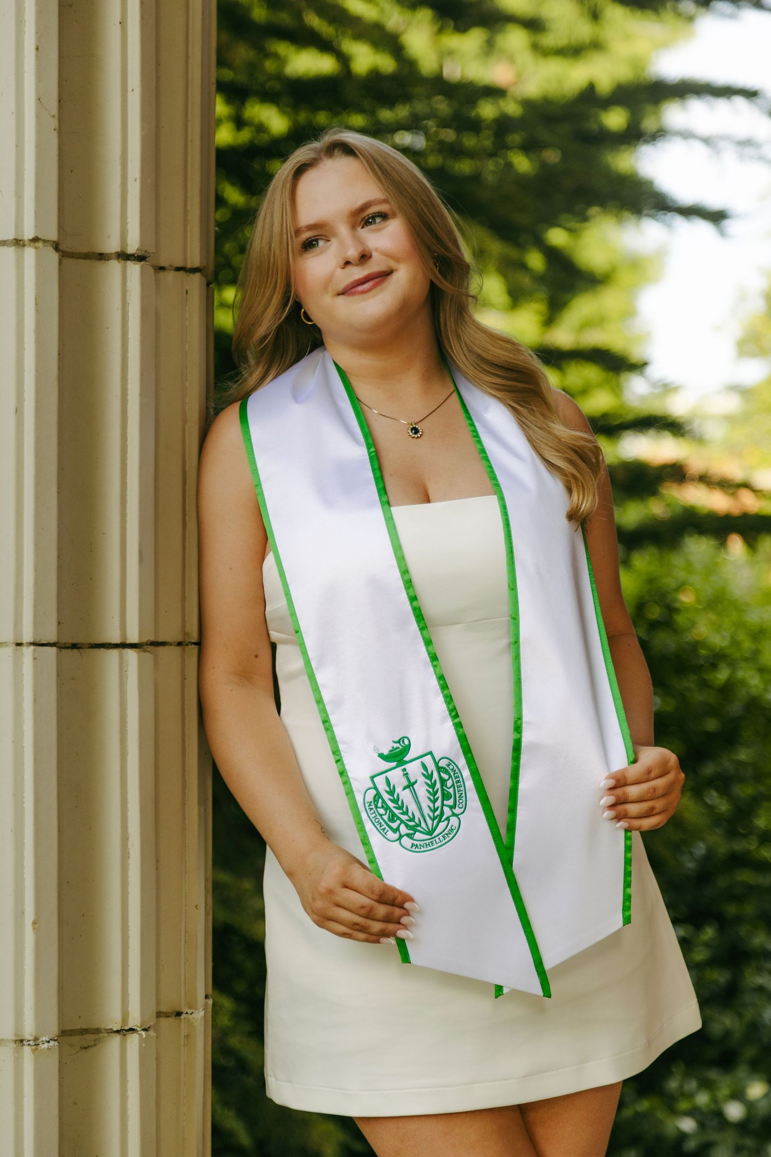 Young woman in white dress wearing a green and white graduation stole, posing by a column outdoors.