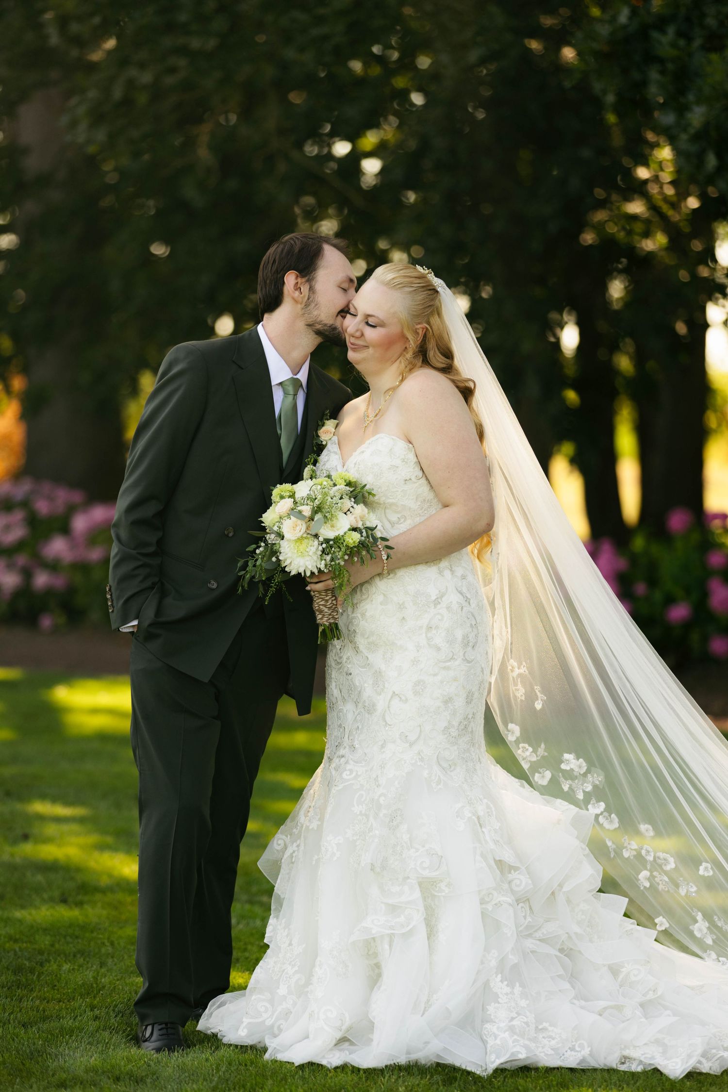 A bride and groom share a romantic moment outdoors, surrounded by lush greenery and pink flowers on their wedding day.