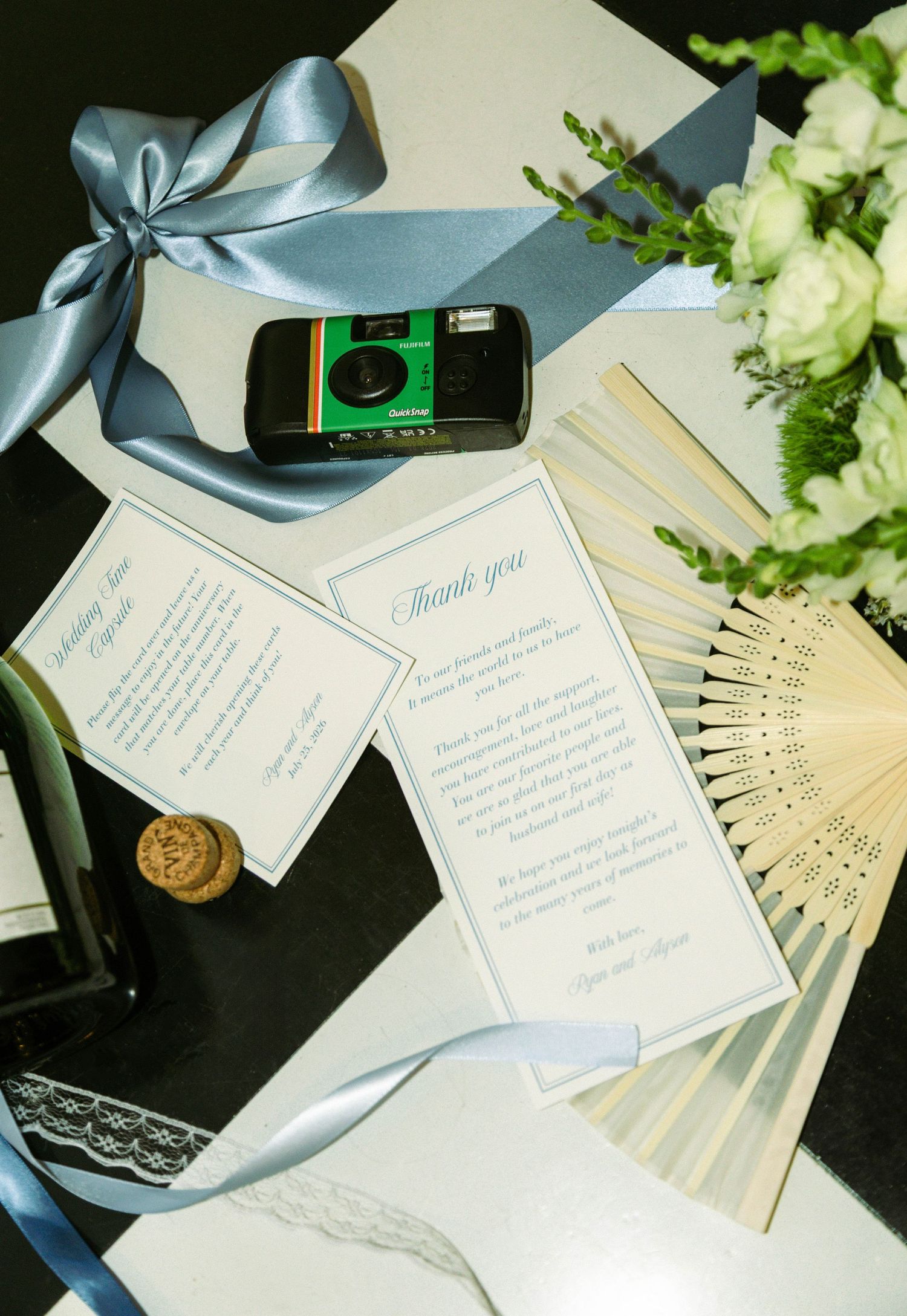 Wedding stationery flat lay with thank you cards, disposable camera, blue ribbon, fan, and white flowers on dark surface.