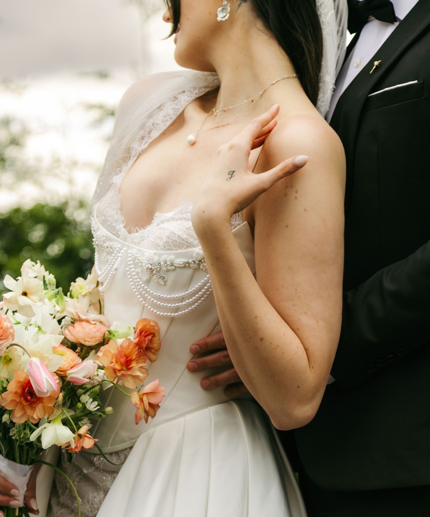 Bride in lace wedding dress holding peach floral bouquet, groom in black tuxedo embracing her shoulders.