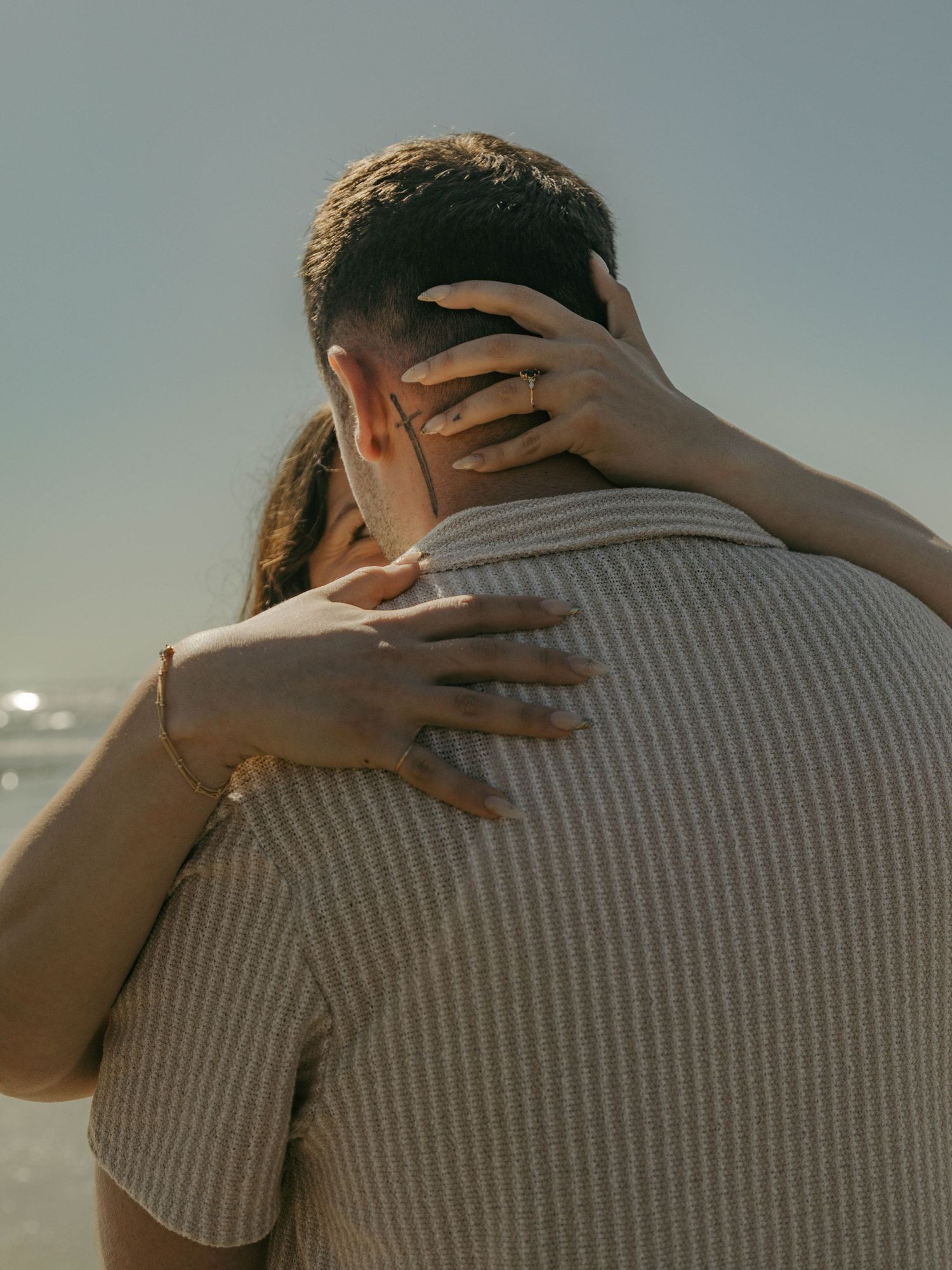 A couple embraces on a beach, woman's hand with ring resting on man's shoulder in warm sunlight.