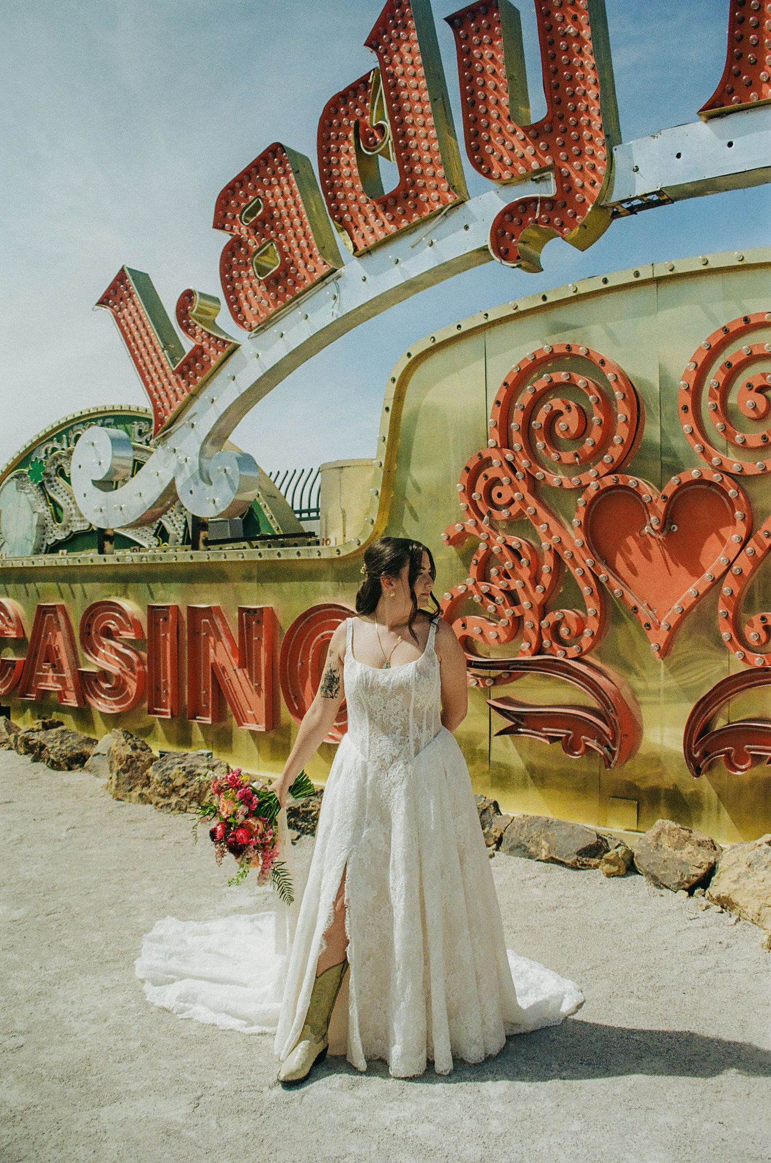 Bride in lace wedding gown poses at the Neon Museum in Las Vegas, holding a bouquet near vintage casino signs.