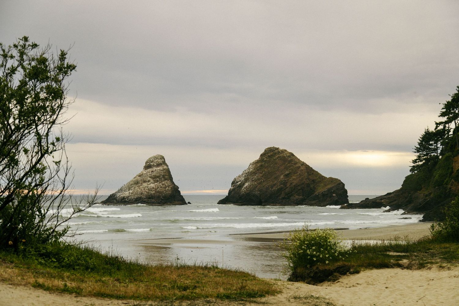 Two large sea stacks rise from a sandy Pacific Northwest beach under overcast skies, framed by coastal trees.