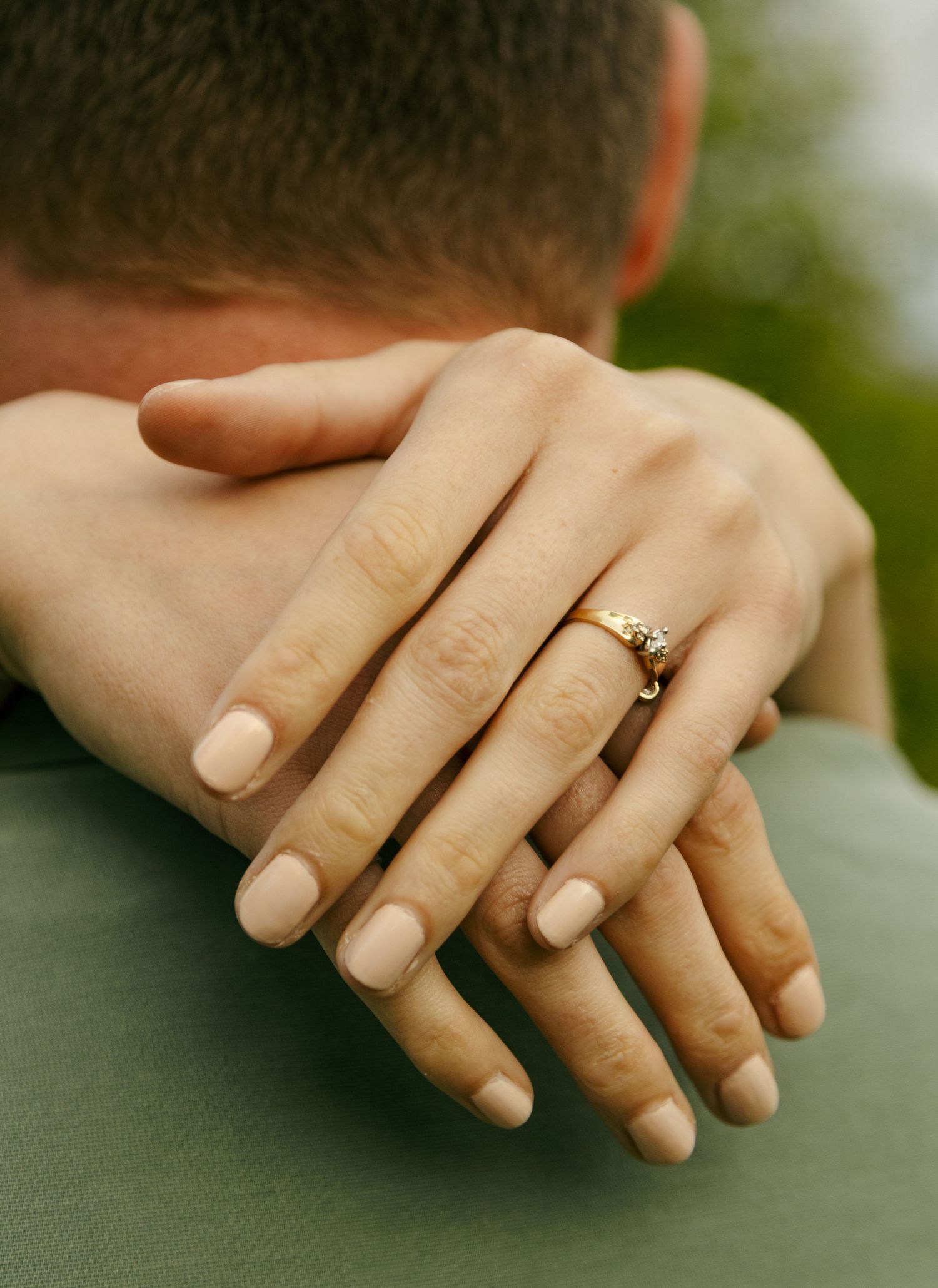 Close-up of a woman's hand with nude nails and gold engagement ring resting on a man's shoulder.