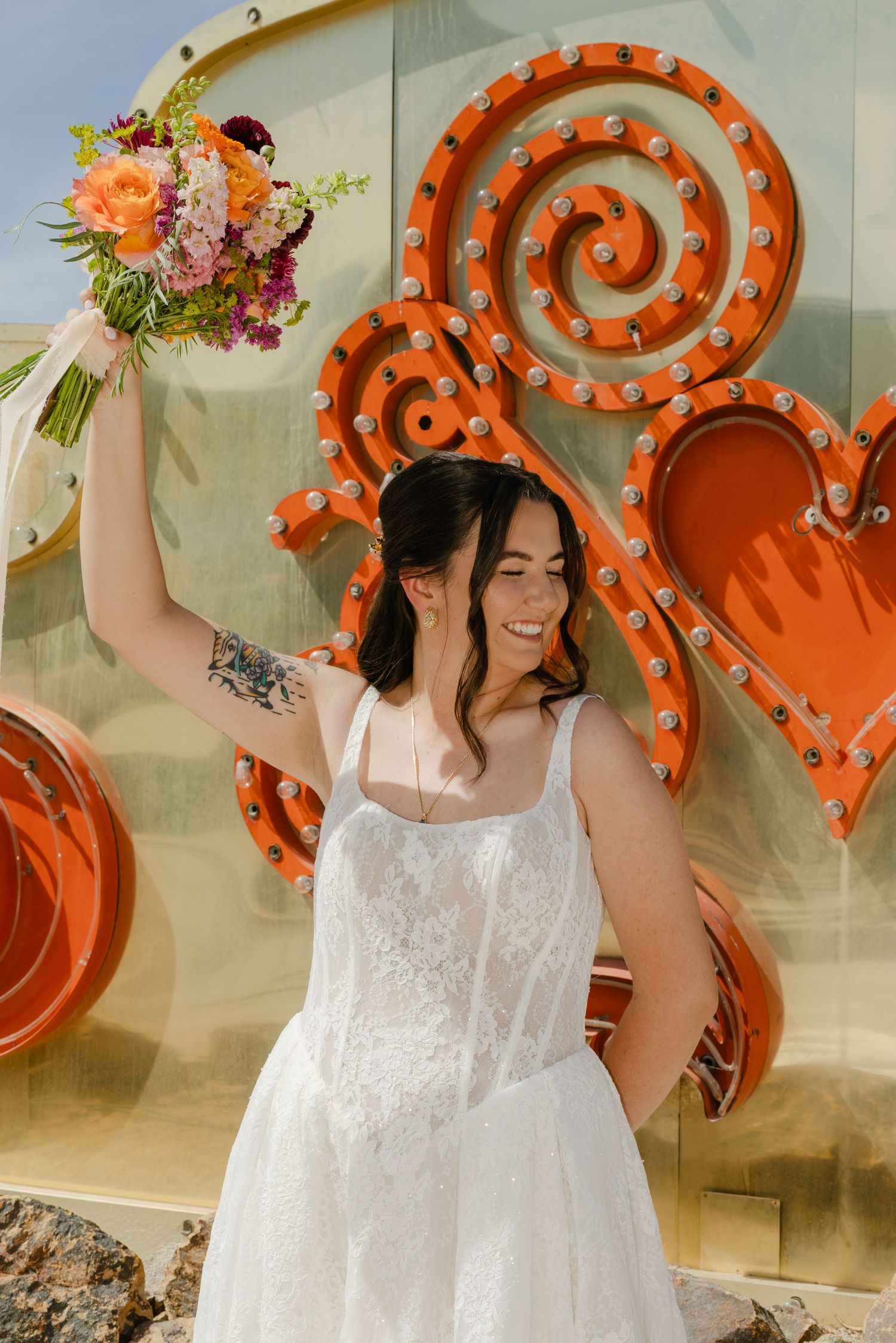 Smiling bride in white lace gown raises colorful bouquet beside vibrant orange neon signs at the Neon Museum Las Vegas.