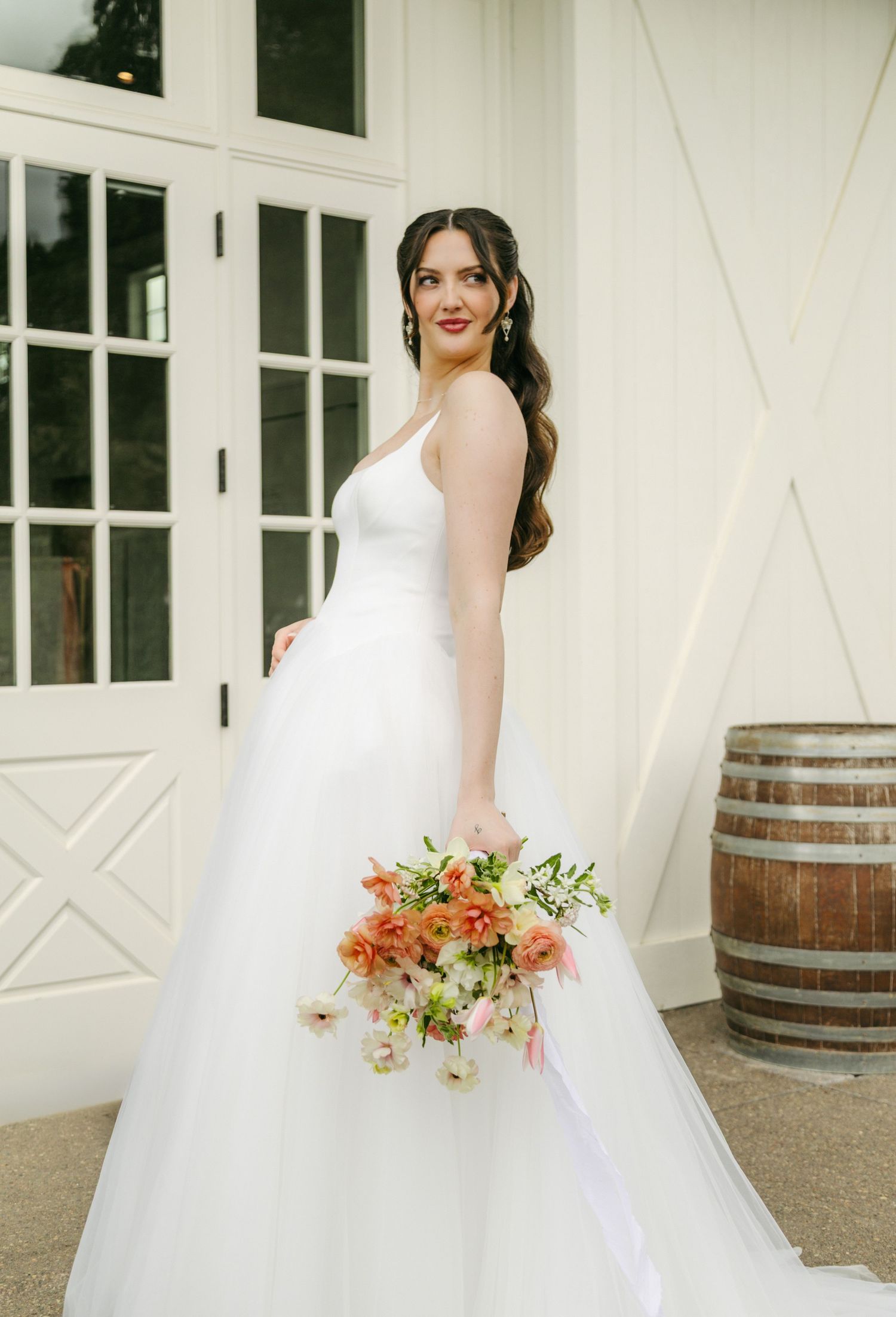 Bride in a white tulle ball gown holding a peach floral bouquet, posing in front of white barn doors.